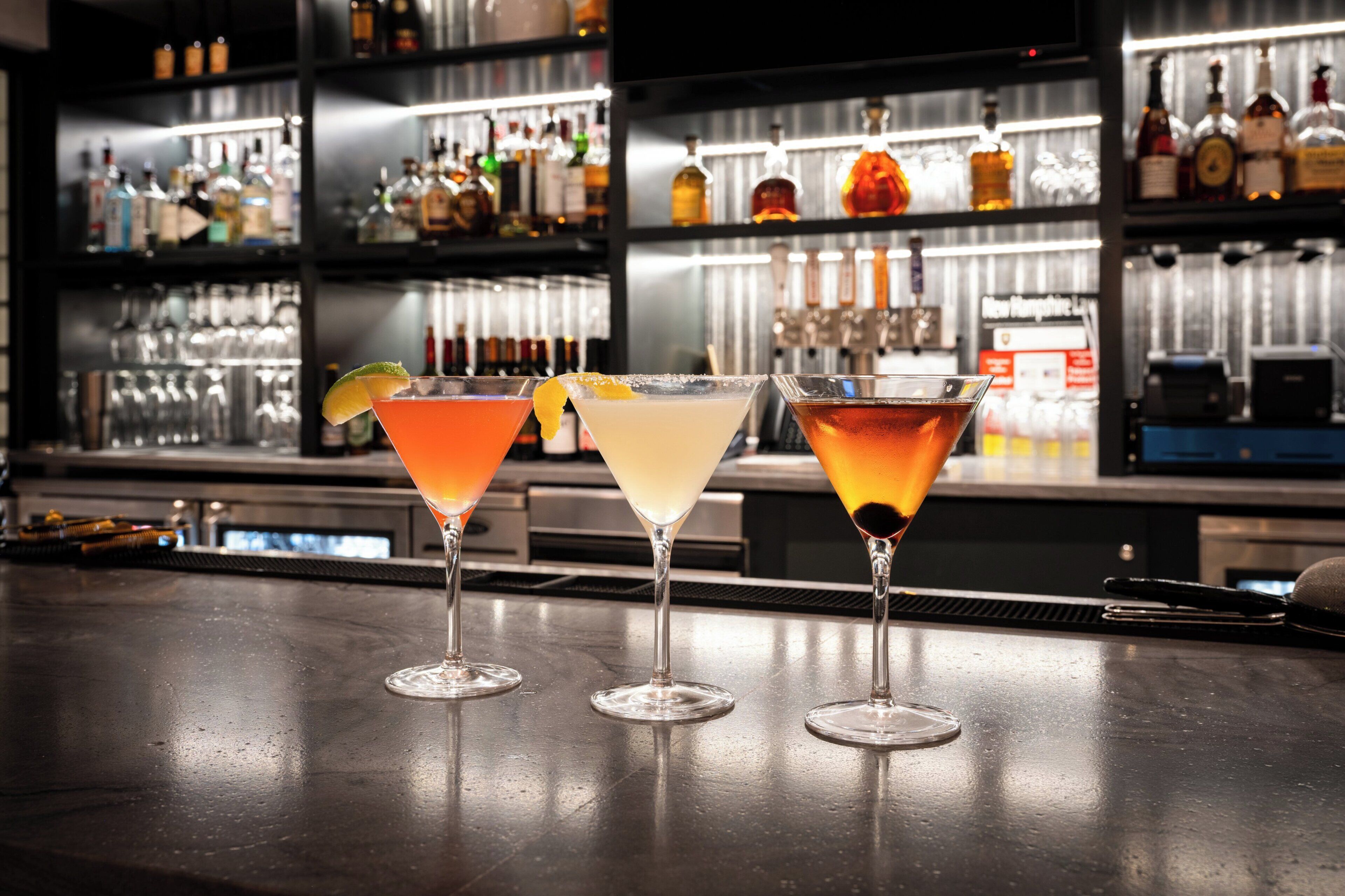 Three martini glasses on a bar counter with a background of shelves stocked with bottles and glasses.