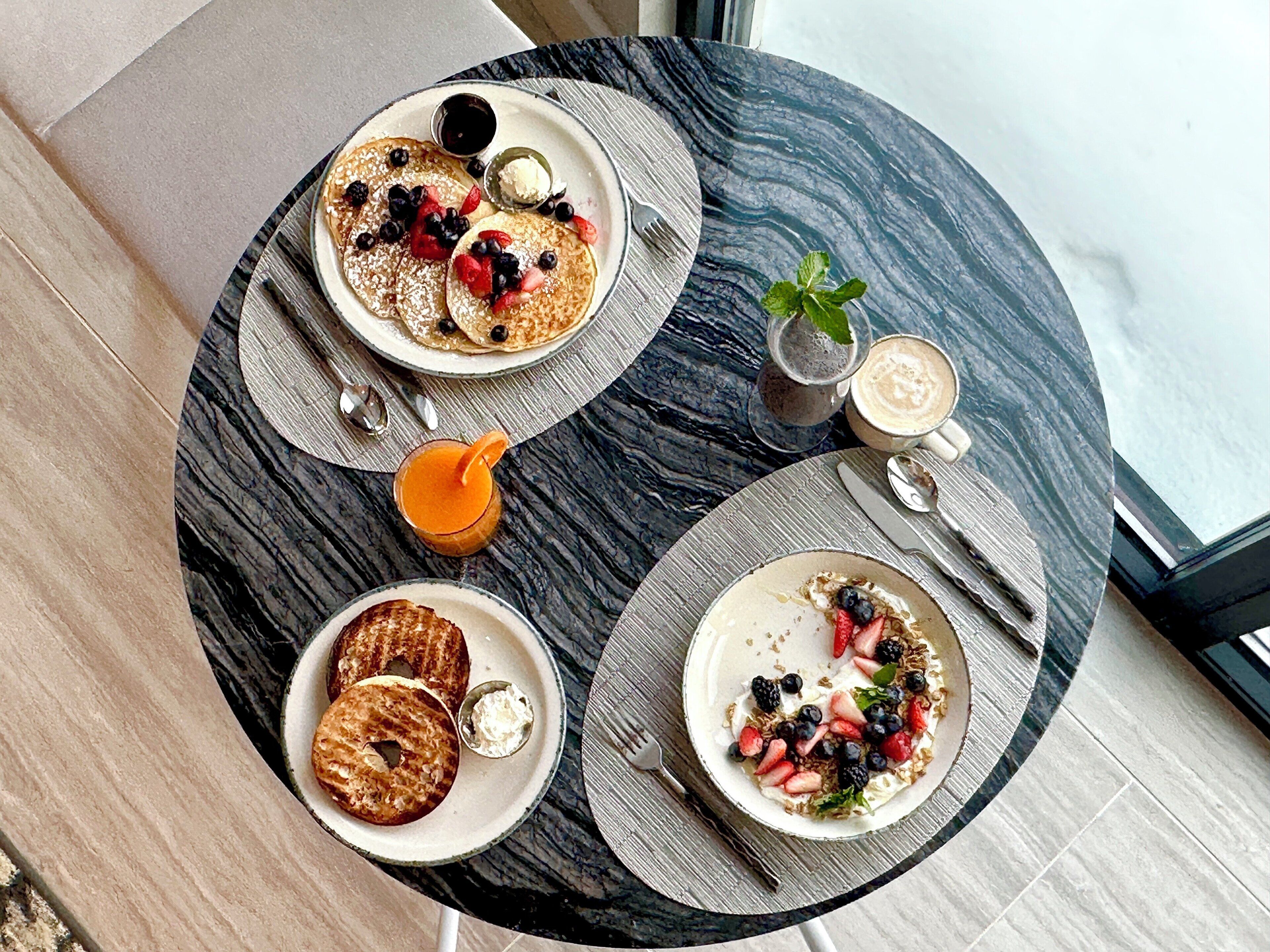 Una escena de desayuno con tortitas, bagels y fruta en platos, un vaso de zumo de naranja y una taza de café sobre una mesa redonda.
