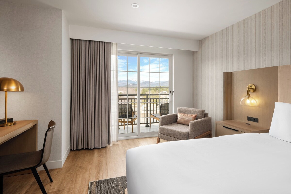 Habitación de hotel con cama, escritorio, silla, mesita de noche, lámpara, luz en la pared y vista a través de un ventanal grande.