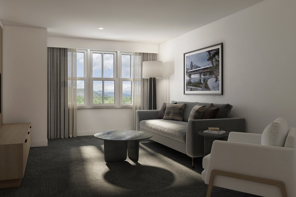 A modern hotel room with a grey sofa, a white armchair, a round coffee table, and a large window offering a view of greenery and mountains.