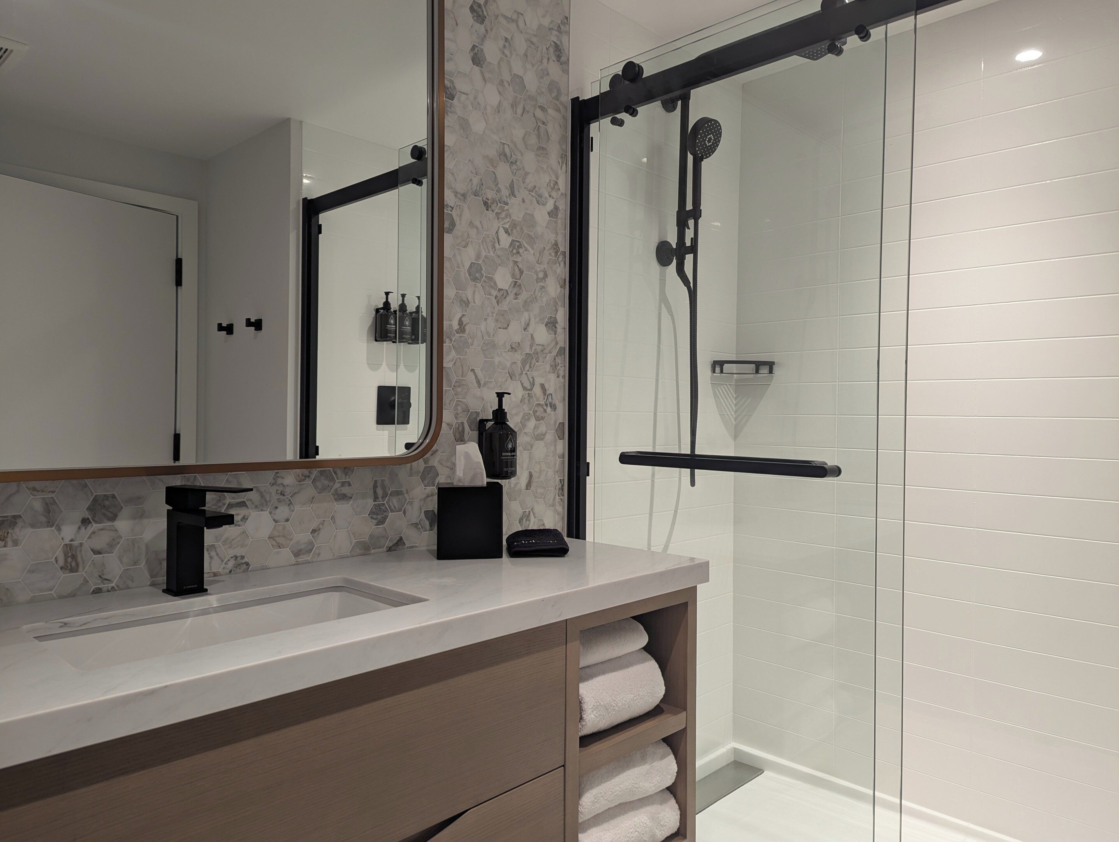 A modern bathroom with a glass shower enclosure, a white sink with a black faucet, and a mirror with a hexagonal tile backsplash.