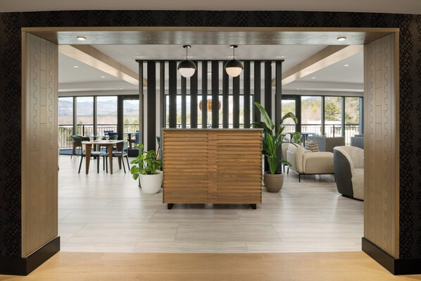 A modern hotel lobby with a wooden reception desk, seating area, large windows, and potted plants.