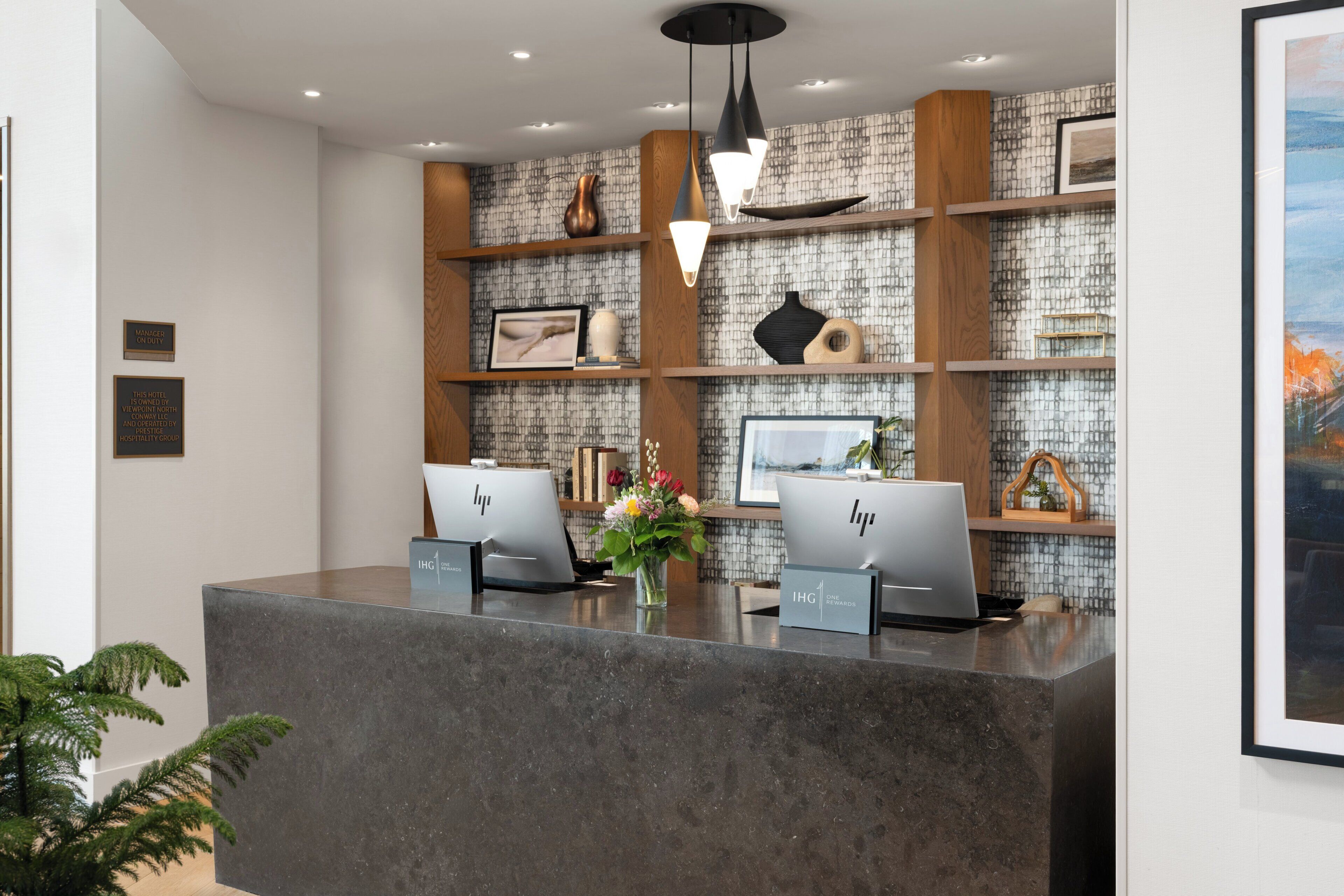 A reception desk with two laptops, a vase of flowers, and a tiled wall with shelves.