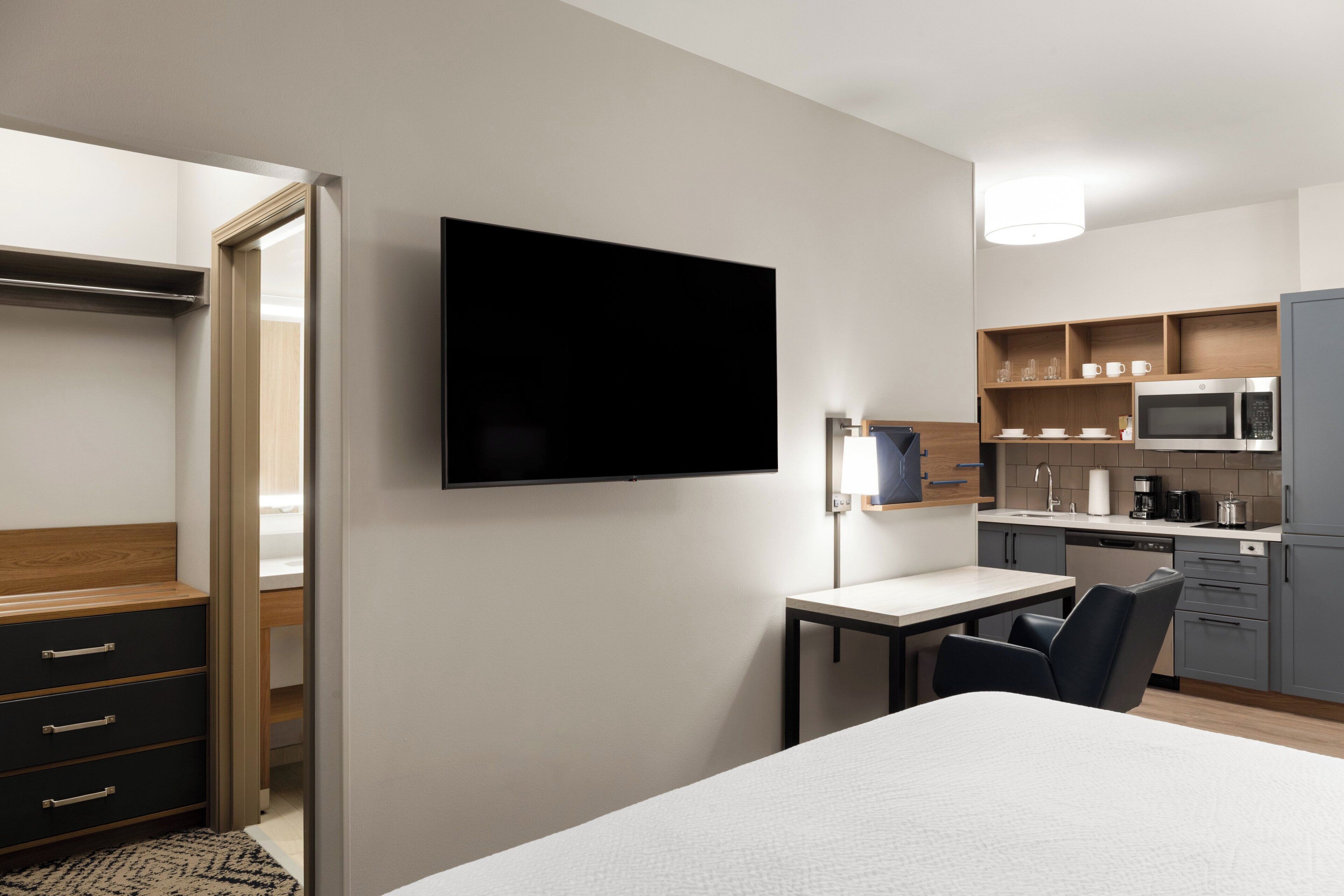 Ett modernt hotellrum med en säng, ett skrivbord, en tv, en garderob och ett litet kök.