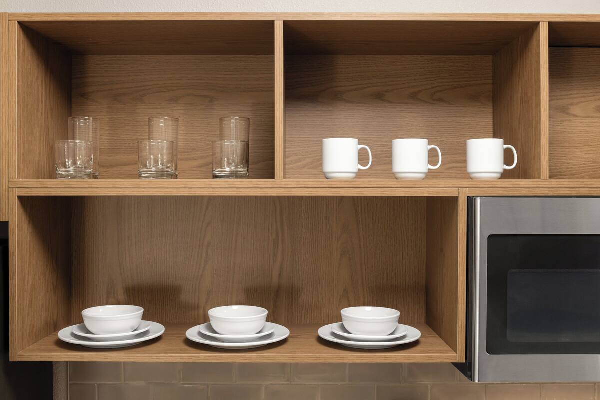 Una cocina con estantes de madera que sostienen vasos transparentes, tazas y recipientes blancos sobre platos.