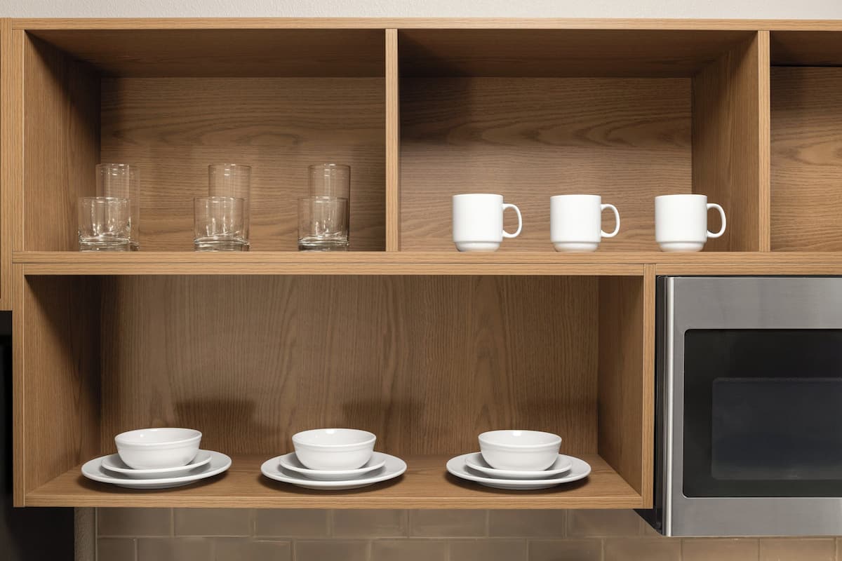 Una cocina con estantes de madera que sostienen vasos transparentes, tazas y recipientes blancos sobre platos.