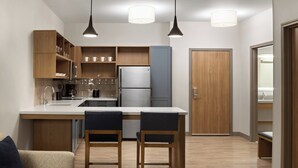Een compacte keuken met een centraal eiland, houten kastjes en een koelkast. Er staan twee barkrukken bij het eiland en een houten deur aan de rechterkant.