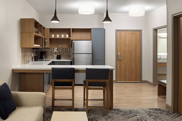 Une cuisine compacte dotée d’un îlot central, de meubles en bois et d’un réfrigérateur. L’îlot est agrémenté de deux tabourets de bar, et une porte en bois se trouve sur le côté droit.