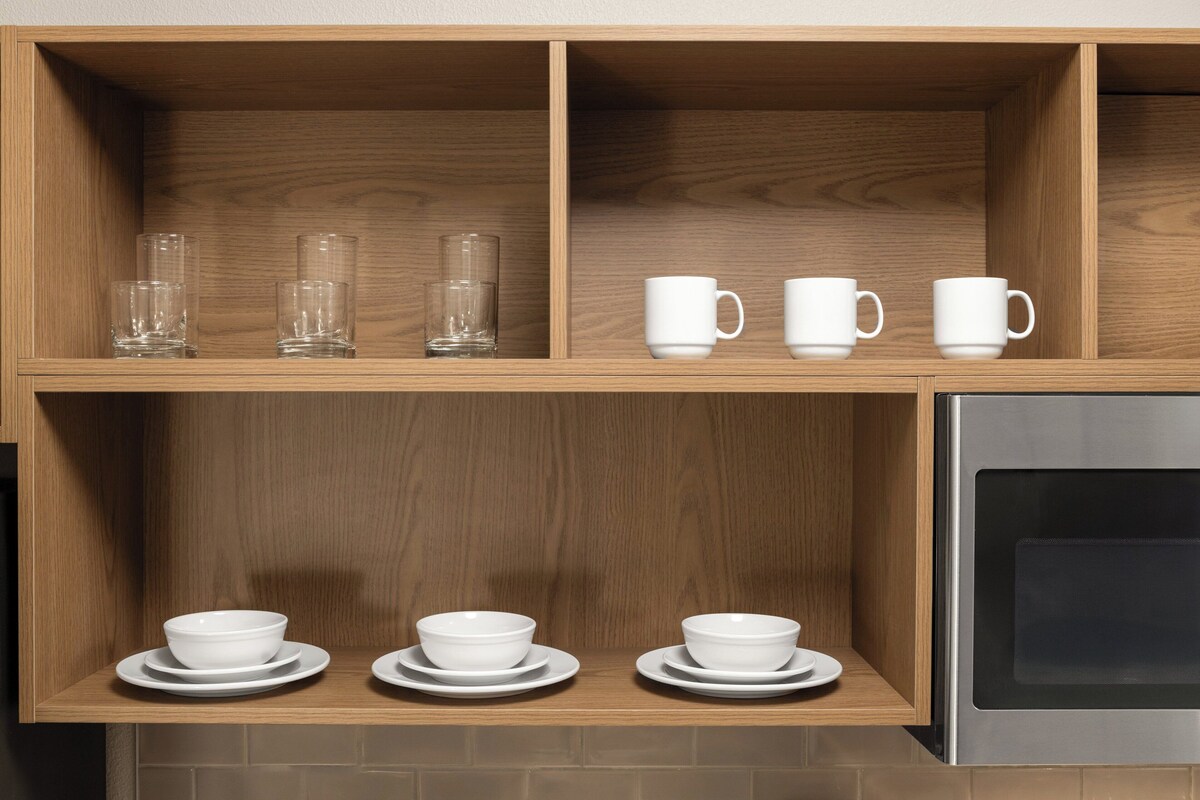 A kitchen with wooden shelves holding clear glasses, white cups, and white bowls on plates.