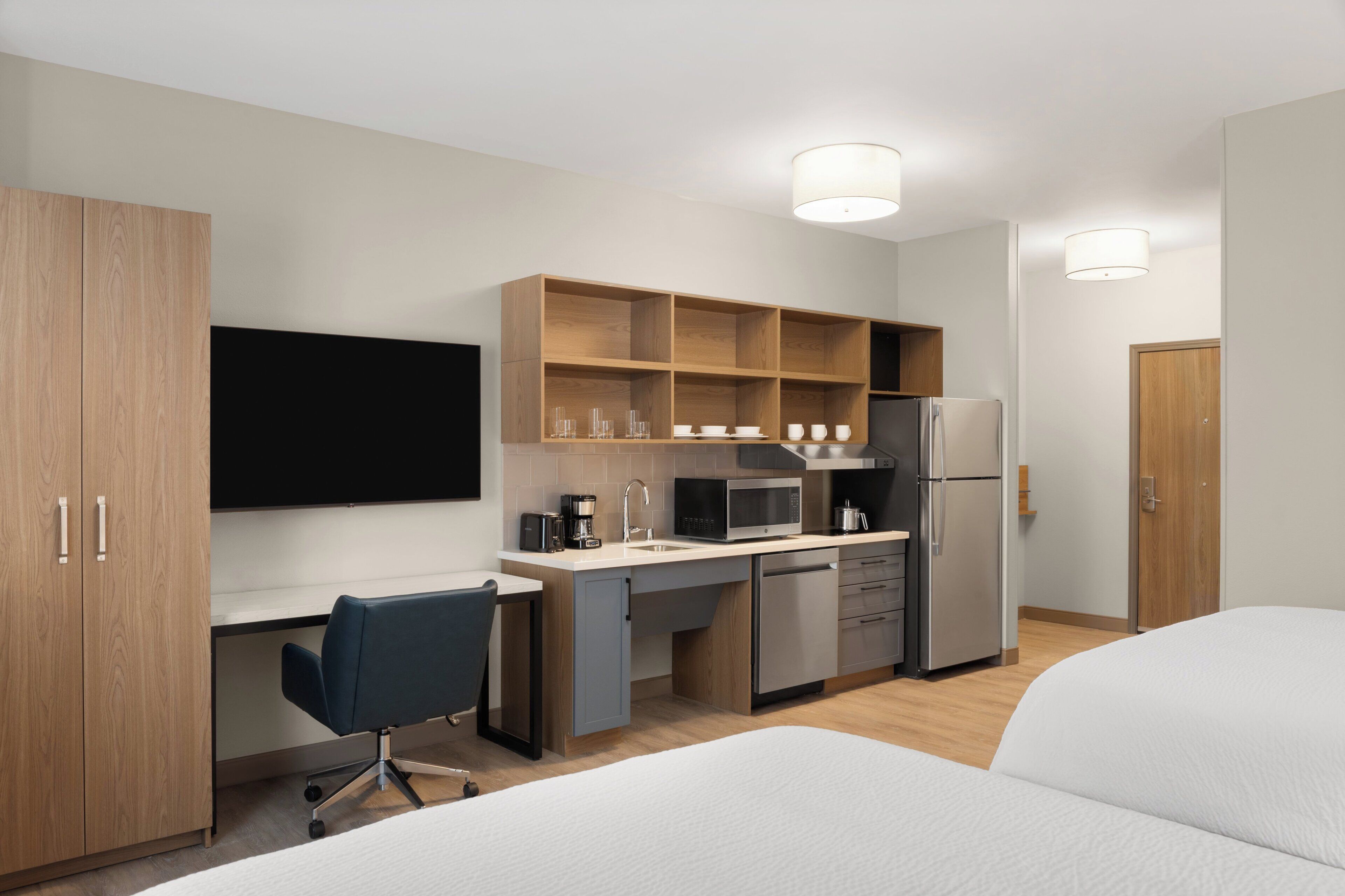 A modern hotel room with a kitchenette, a bed, a desk, and a chair.