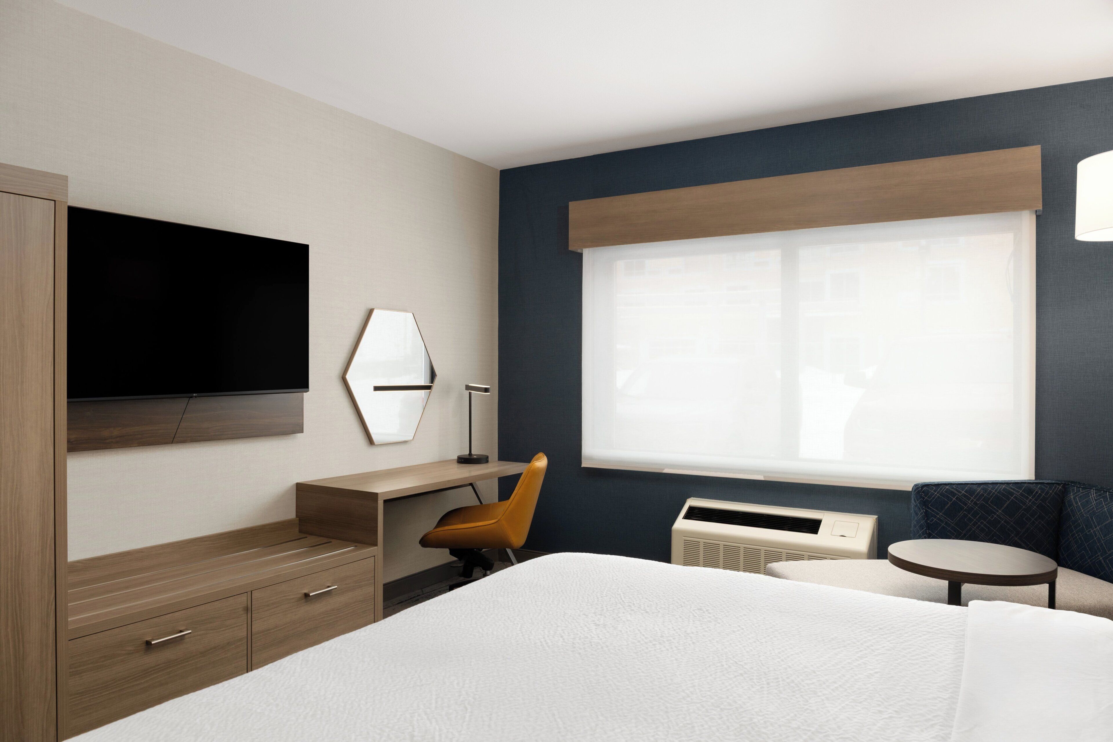 A hotel room with a bed, a desk, a chair, a TV, a window, and a small table.