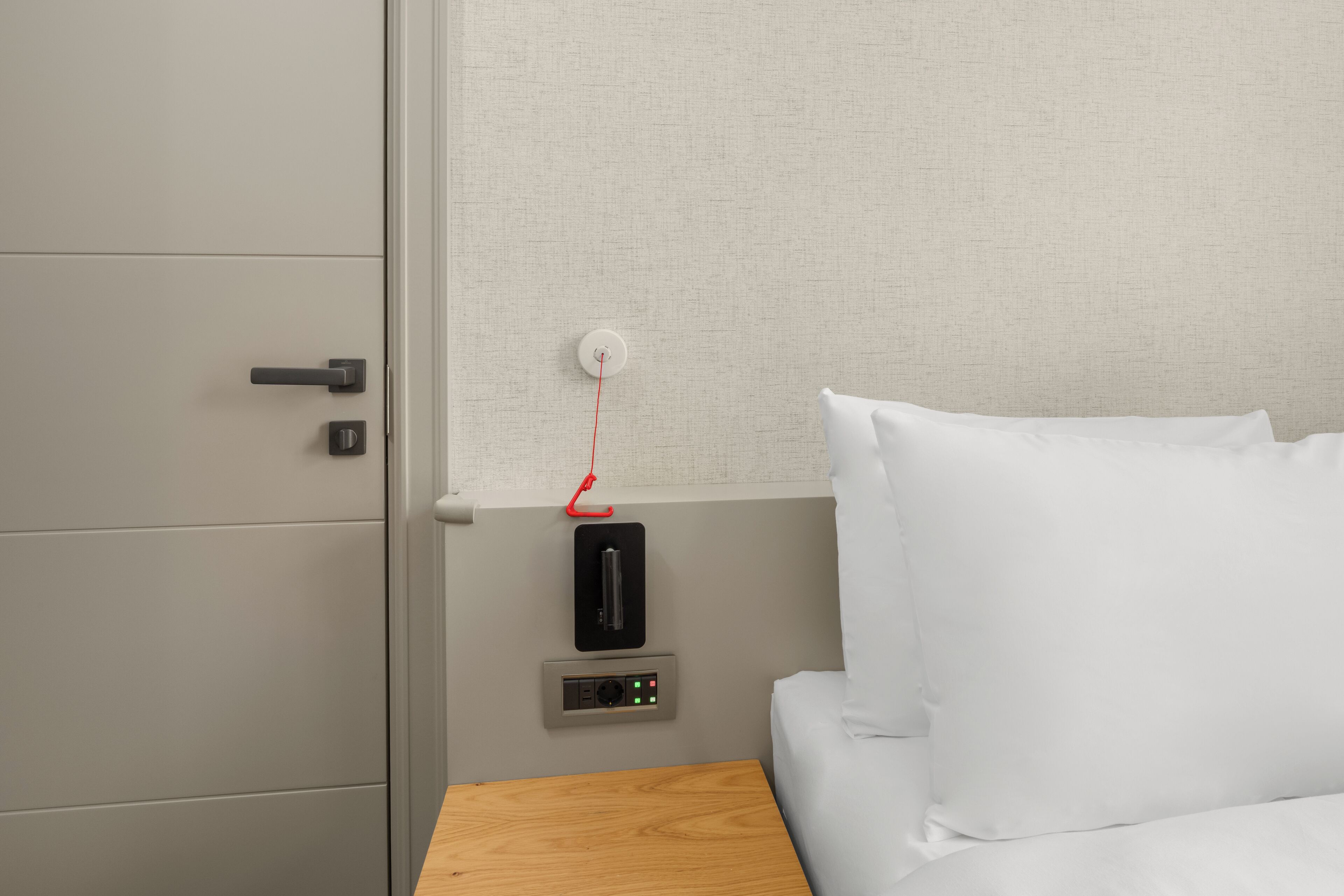 A hotel room with a wooden nightstand, a bed with white bedding, a grey wardrobe, and a wall-mounted control panel.