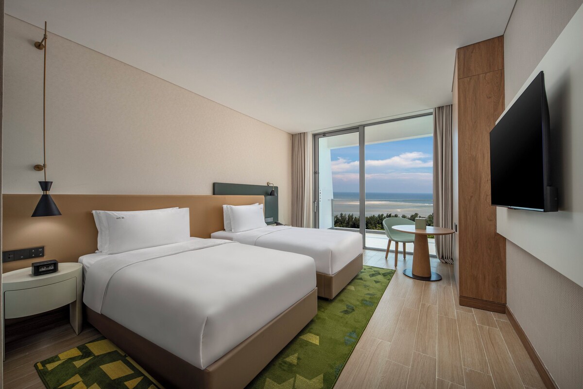 Habitación de hotel con dos camas, televisión, una mesita y vista al océano.