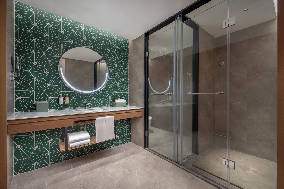 Un baño moderno con un revestimiento de azulejos geométricos, un espejo redondo, un mueble de tocador de madera, una cabina de ducha de vidrio y un perchero para toallas.