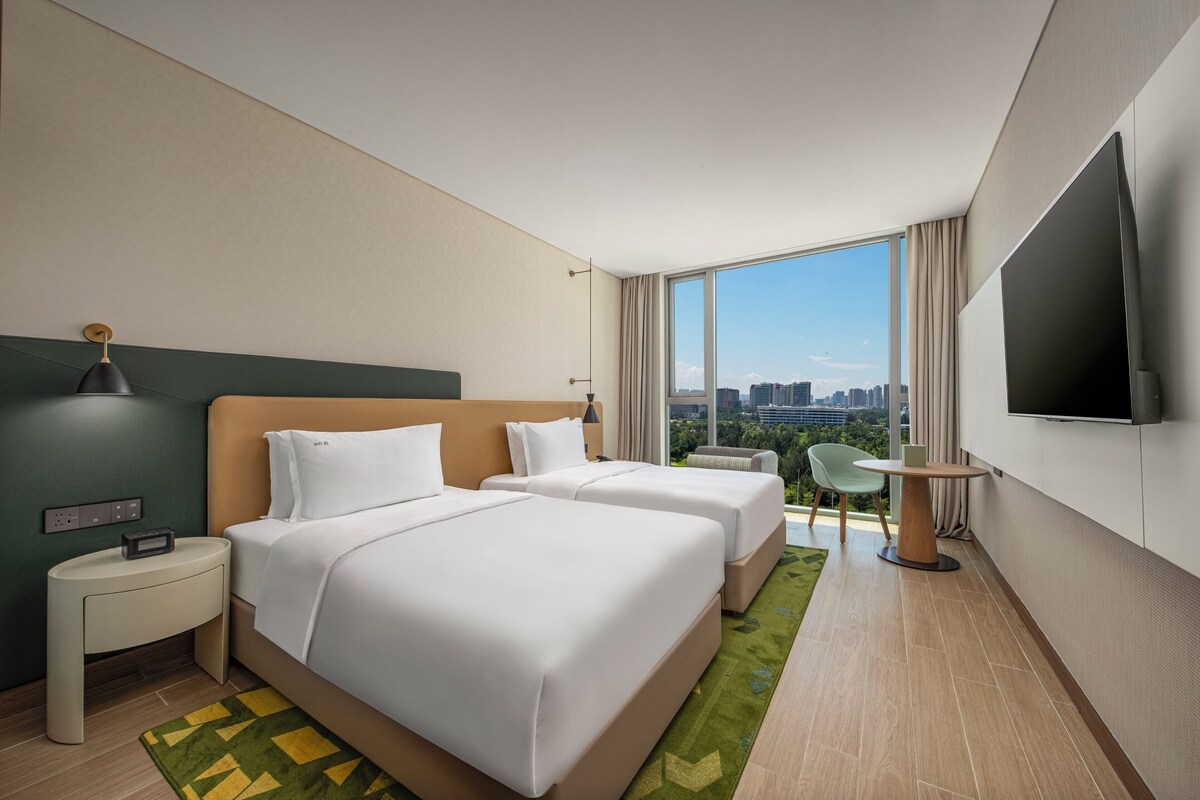 Habitación de hotel con dos camas, televisión, escritorio y vistas a la ciudad.