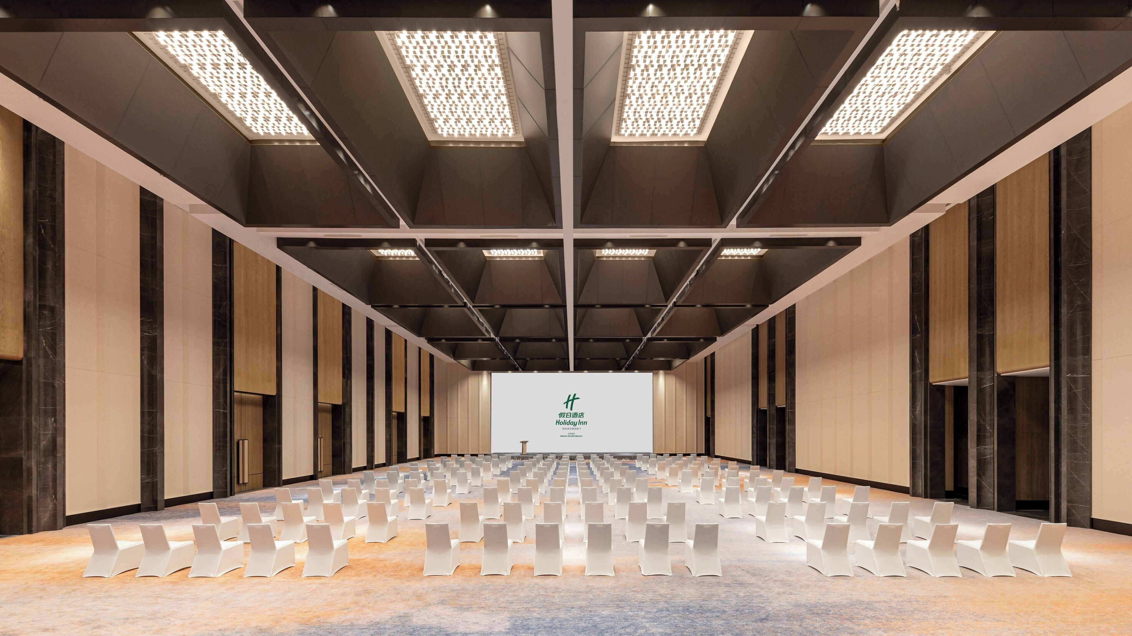 A hotel conference room with rows of chairs, a large screen at the front, and a modern ceiling design.