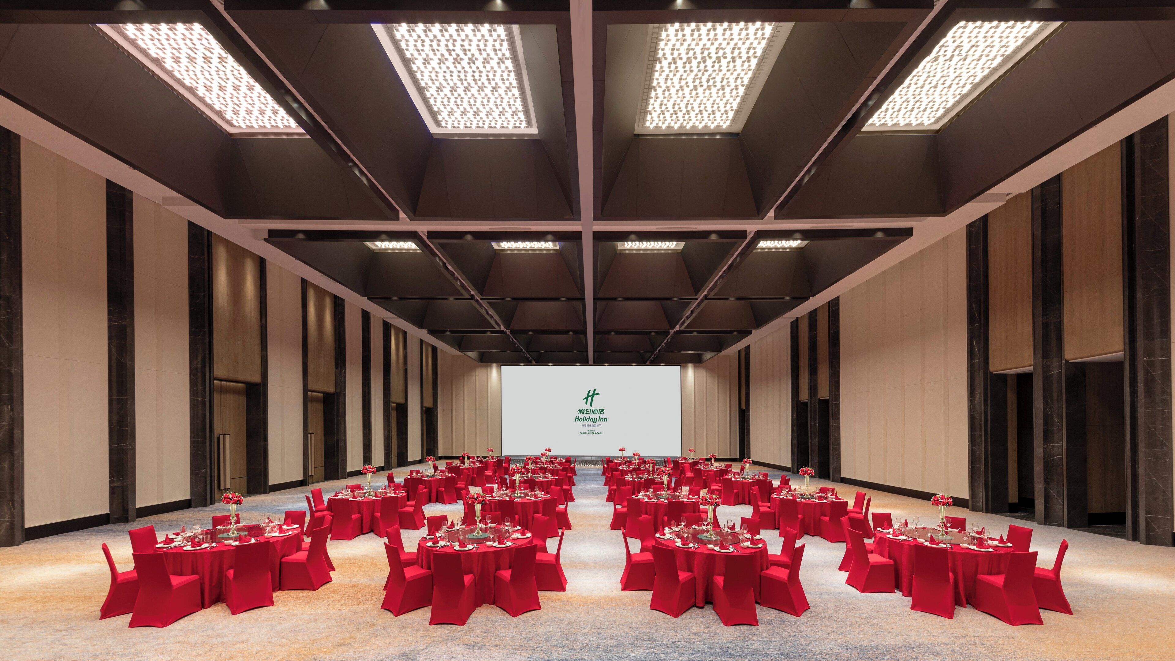 Un salón de conferencias con filas de mesas dispuestas para una reunión, una pantalla grande al frente y un diseño de techo moderno.