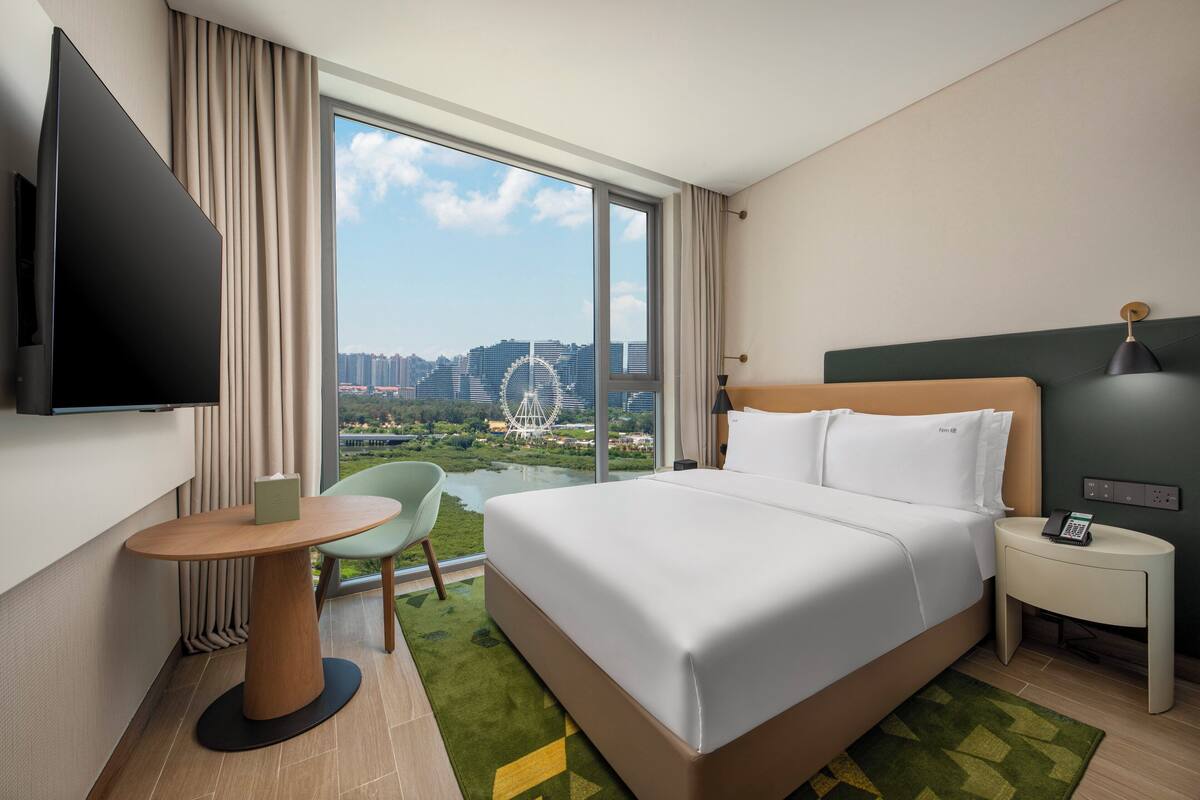 Habitación de hotel con una cama grande, un televisor montado en la pared, una mesita, una silla y vista a una noria a través de un ventanal amplio.