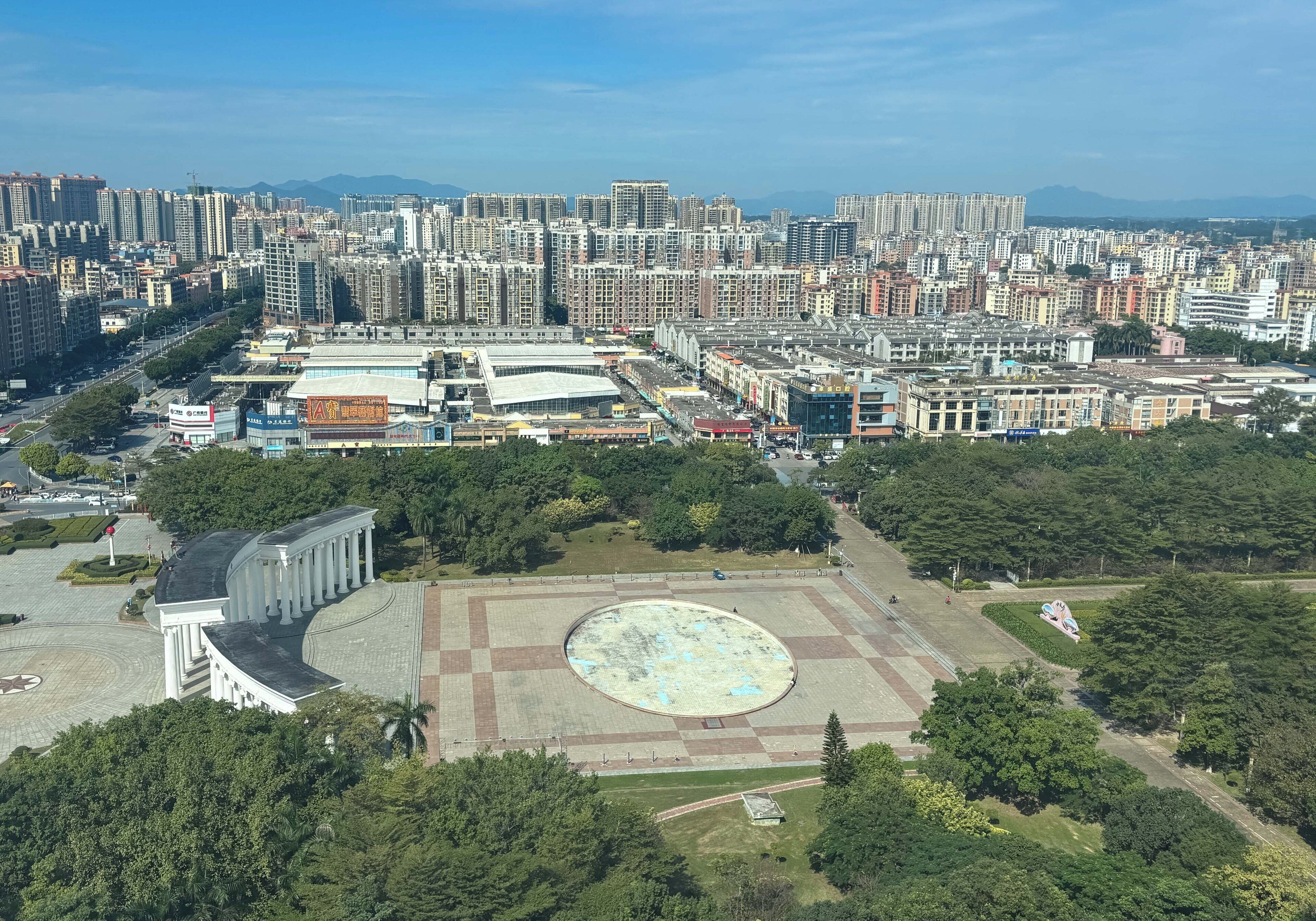 Paisagem urbana com edifícios altos, uma grande praça aberta e áreas verdes ao redor.