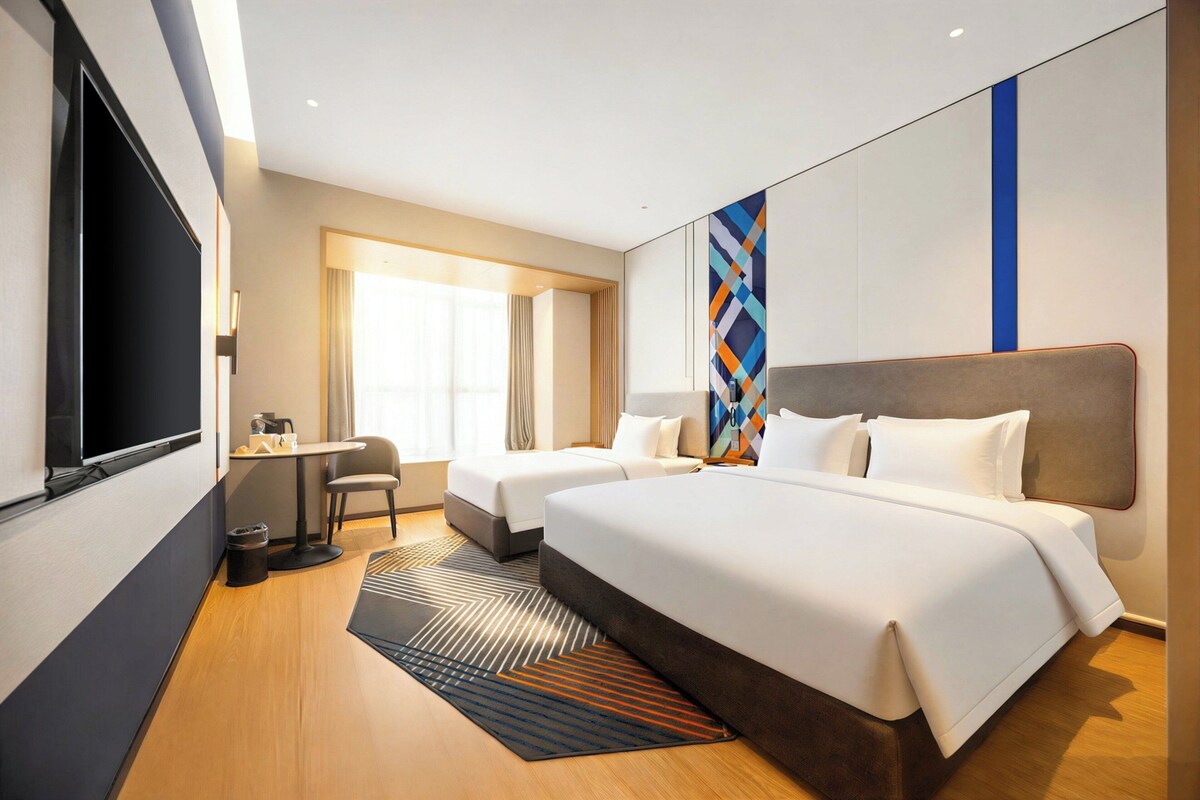 Ein Hotelzimmer mit zwei Betten, einem Schreibtisch, einem Stuhl, einem Fernseher und einem großen Fenster.