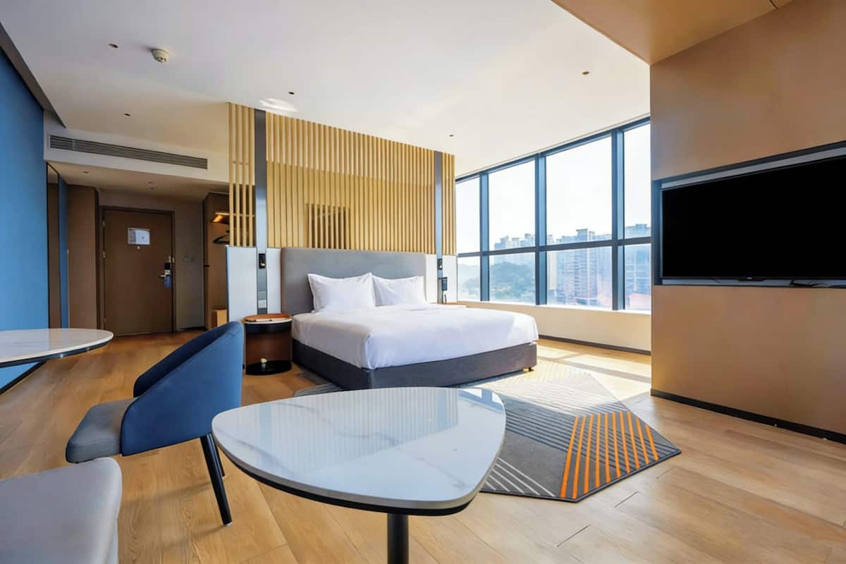 Ein modernes Hotelzimmer mit einem großen Bett, einem Flachbildfernseher, einem Esstisch und Blick auf die Stadt.