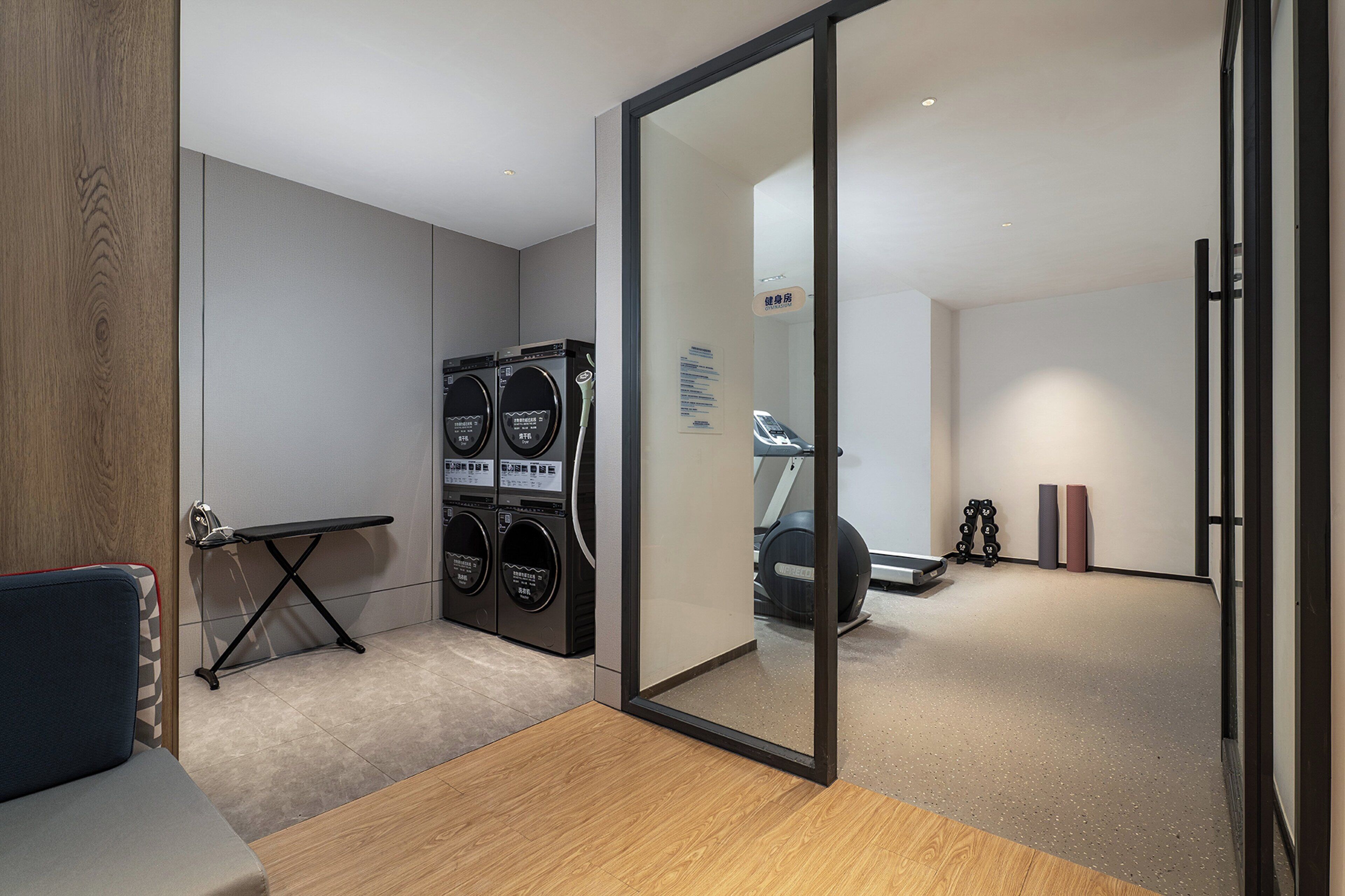 A modern room with a wooden floor, a seating area, a treadmill, and a set of stacked laundry machines.