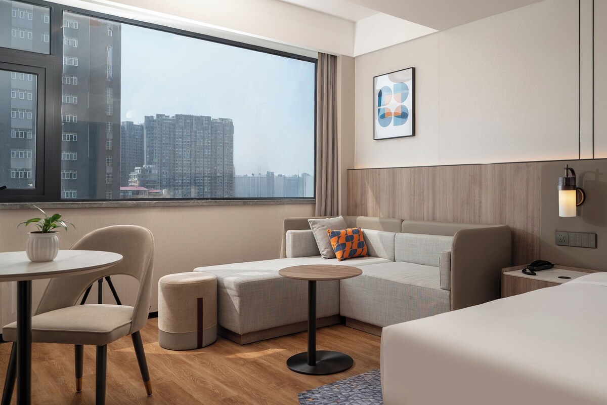 A modern hotel room with a large window, a sofa, a small table, a chair, and a bed.
