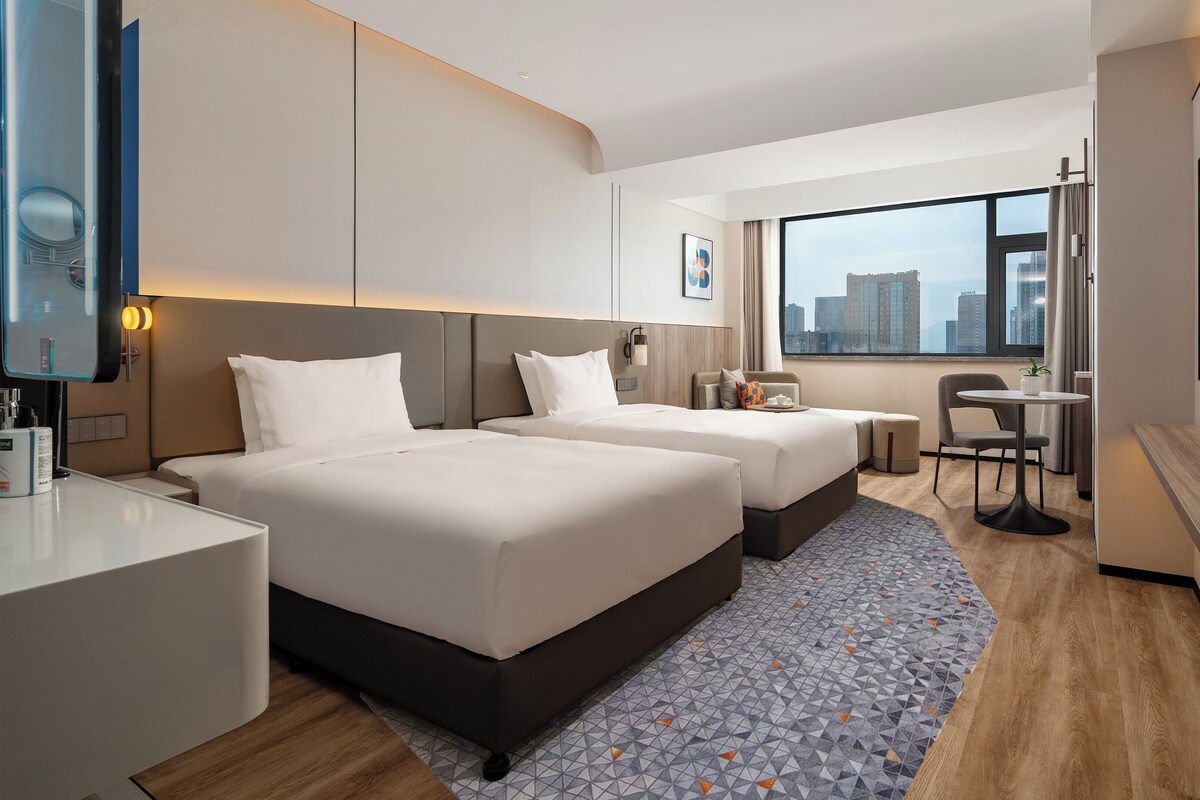A modern hotel room with two beds, a small table, and a seating area.