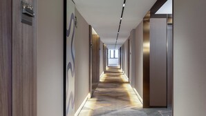 Un couloir d’hôtel moderne, avec un tapis à motifs, des murs en bois et recouverts de tissu, et un long couloir qui mène à un espace lumineux à l’extrémité.