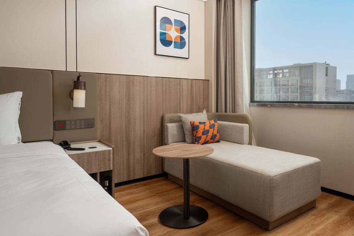 A hotel room with a bed, a small sofa, a round table, and a view of the cityscape through the window.