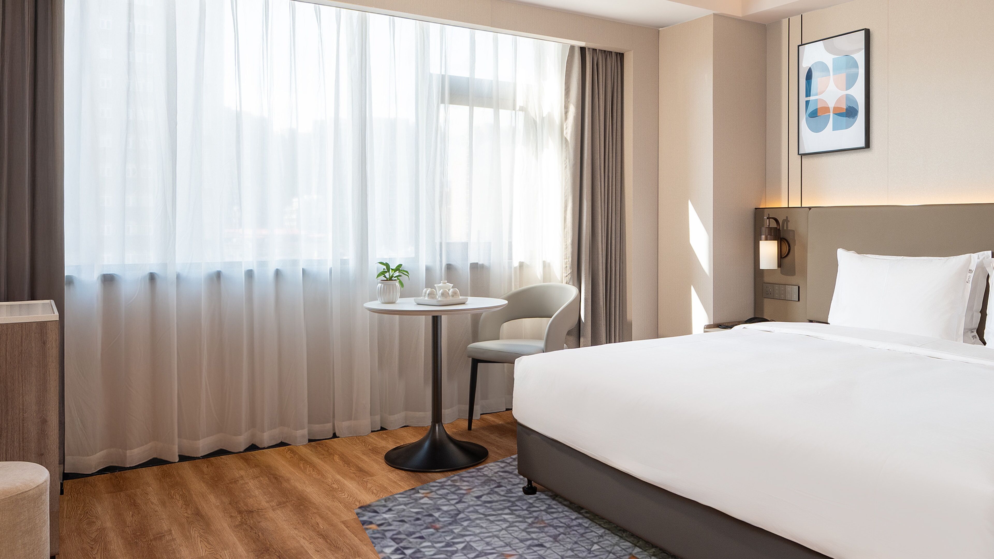 A hotel room with a large bed, a small round table with a plant, a chair, and a wall-mounted light fixture.