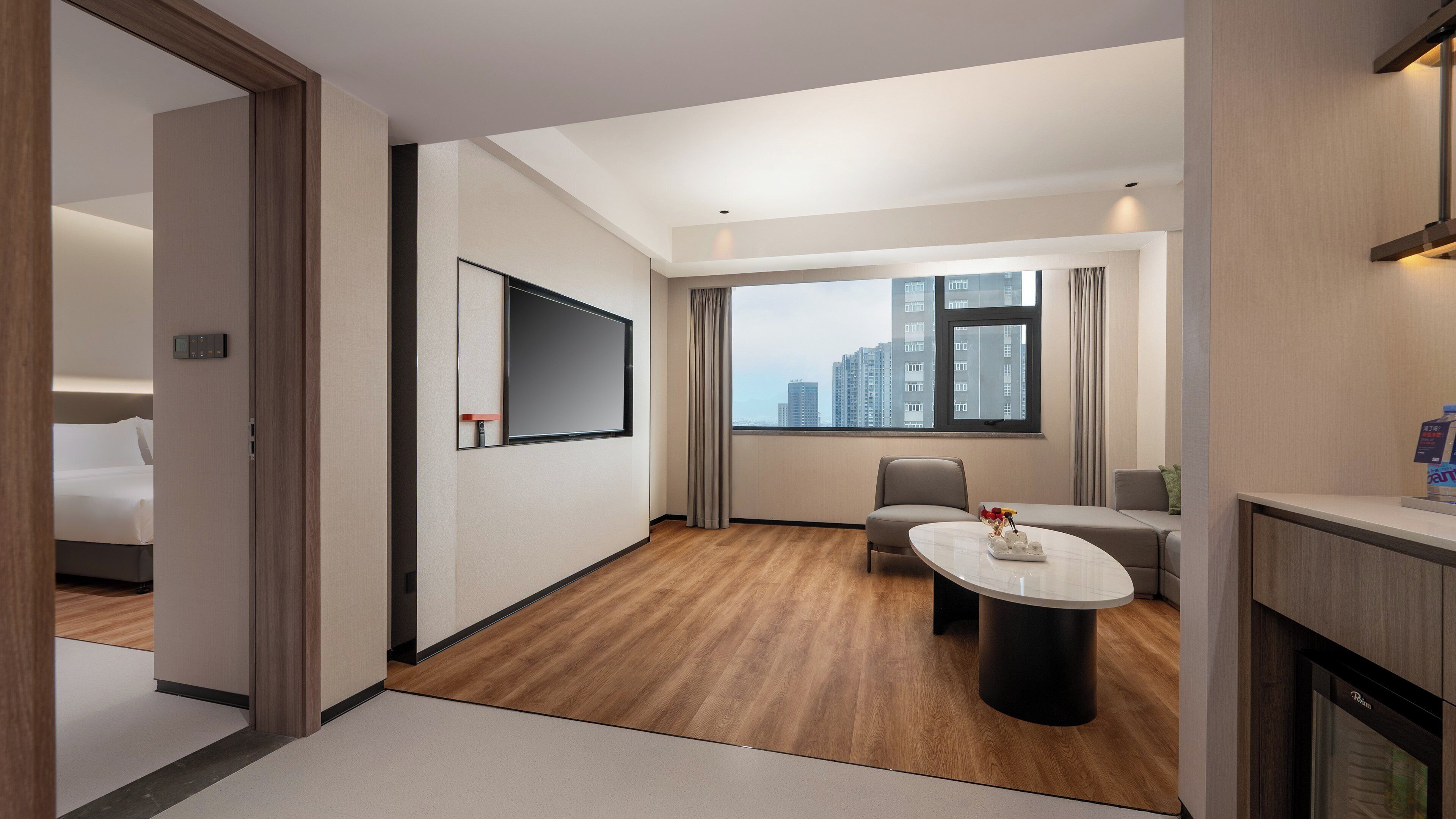 A modern hotel room with a wooden floor, a flat-screen TV mounted on the wall, a dining table with a vase of flowers, and a view of tall buildings through the window.