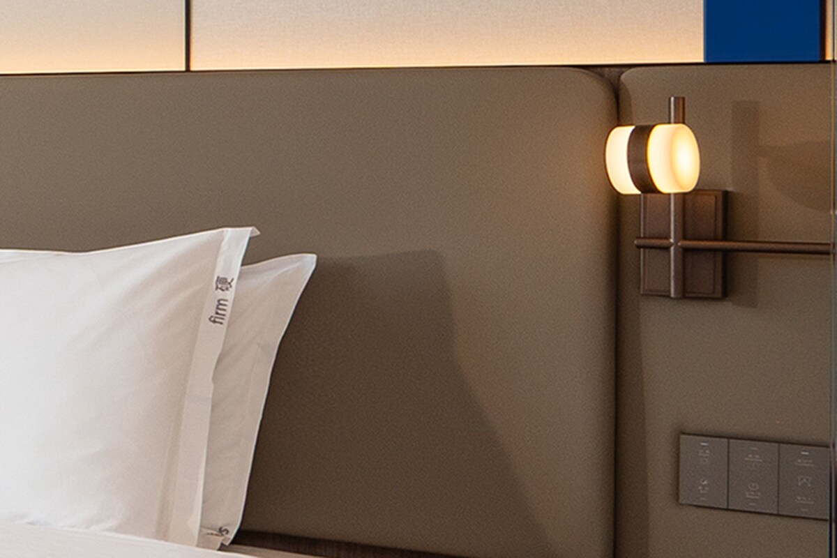 A hotel room with a bed, a headboard, and a wall-mounted light fixture.