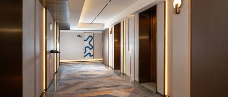 Un pasillo moderno de hotel con alfombra estampada, luminarias fijadas a la pared y una gran obra de arte abstracta en la pared.