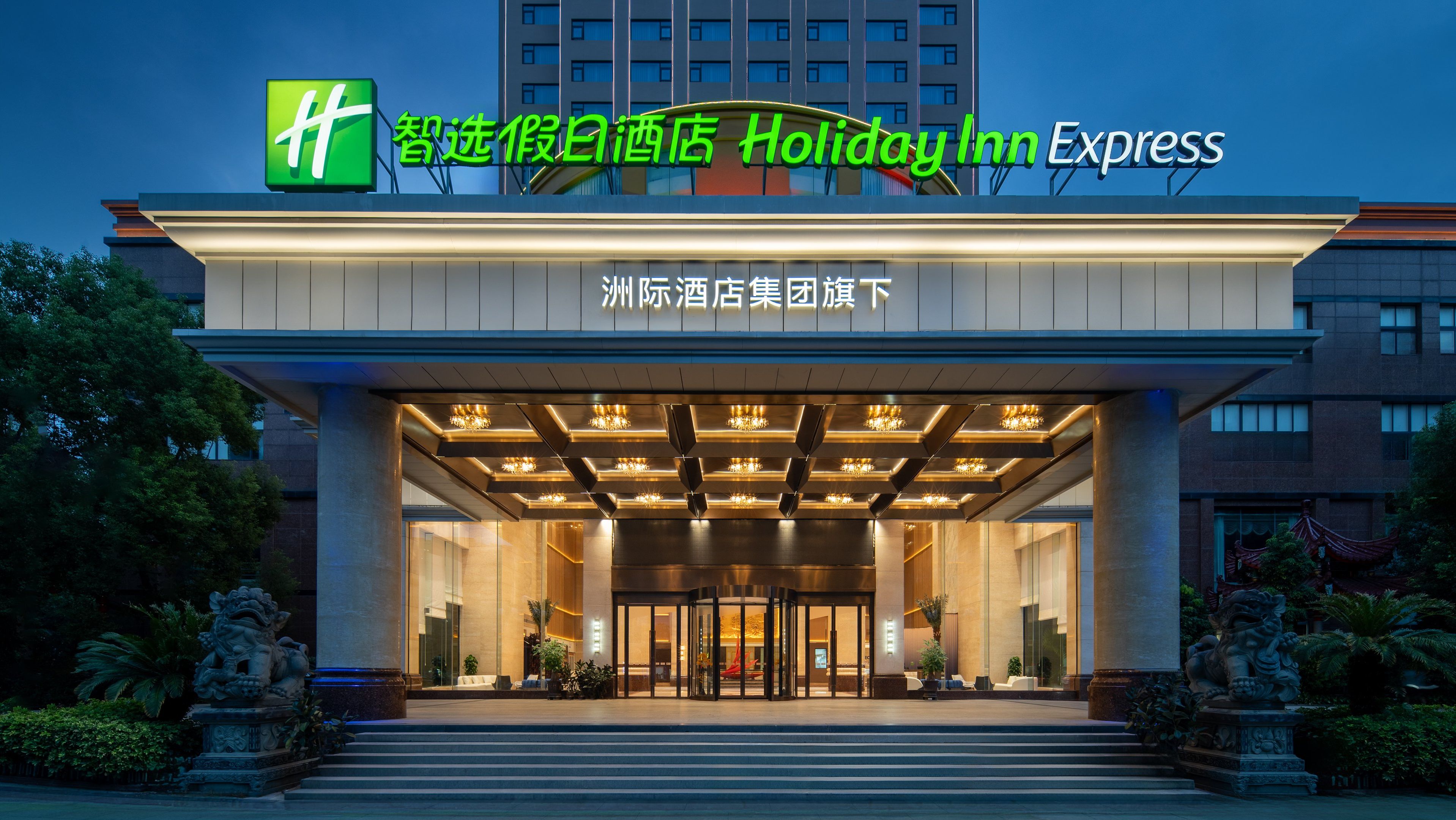 Entrada de un hotel Holiday Inn Express con diseño moderno, destacando un amplio frente de vidrio y un área de acceso cubierta con iluminación decorativa.