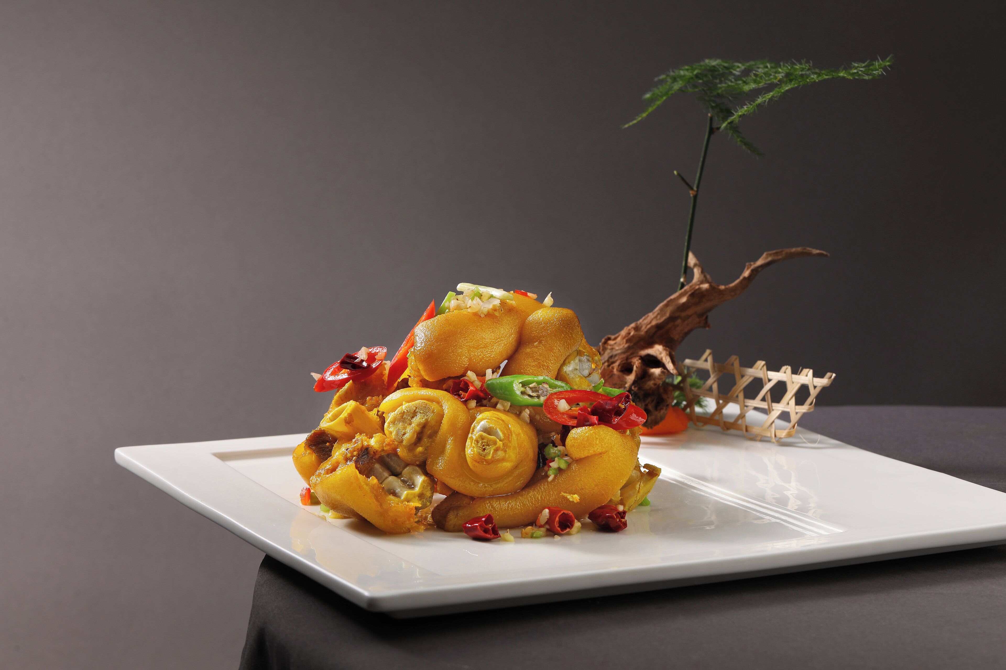 A square white plate with a food arrangement featuring yellow and orange elements, garnished with red and green peppers, and a small plant in the background.