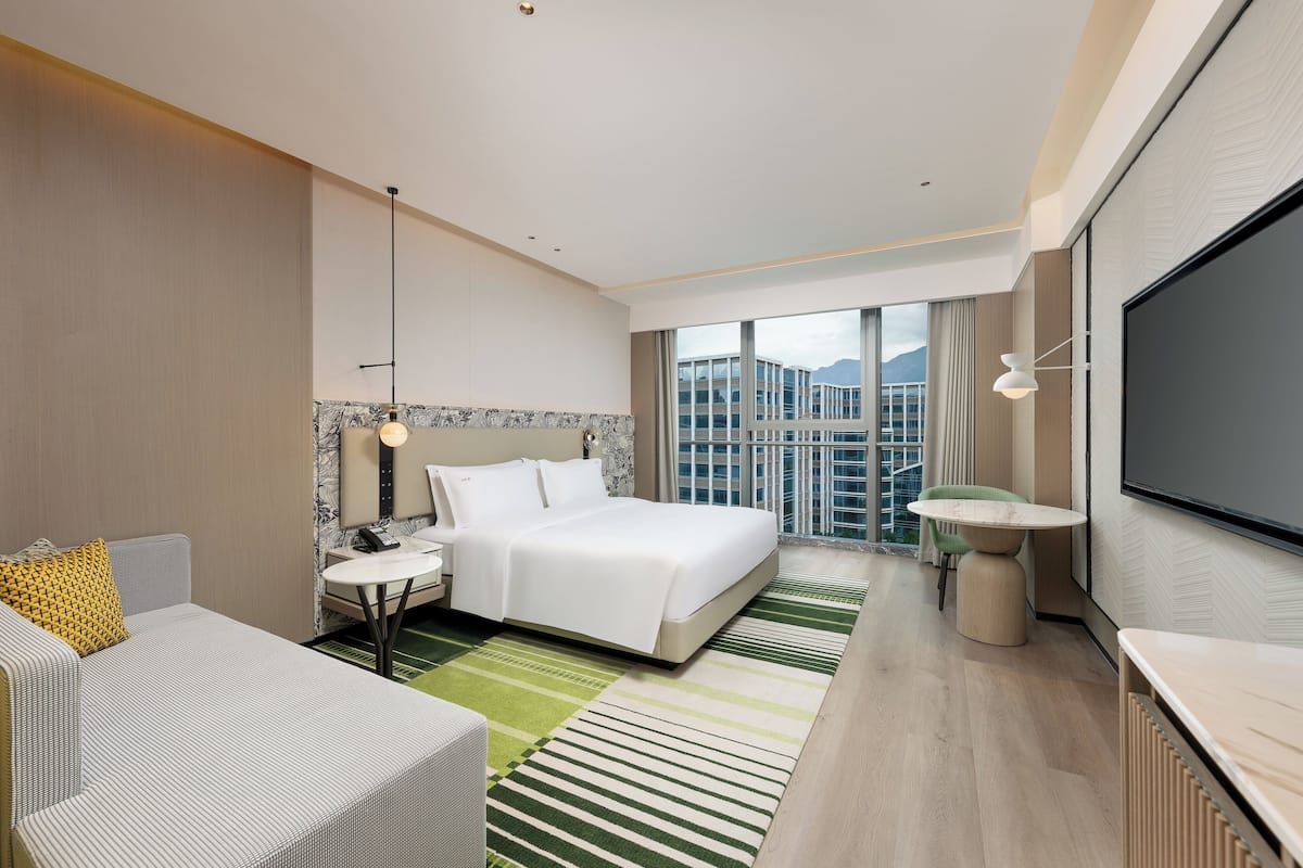 Ein modernes Hotelzimmer mit einem großen Bett, einer Sitzecke mit Tisch, einem Fernseher und Blick auf eine Stadtlandschaft.