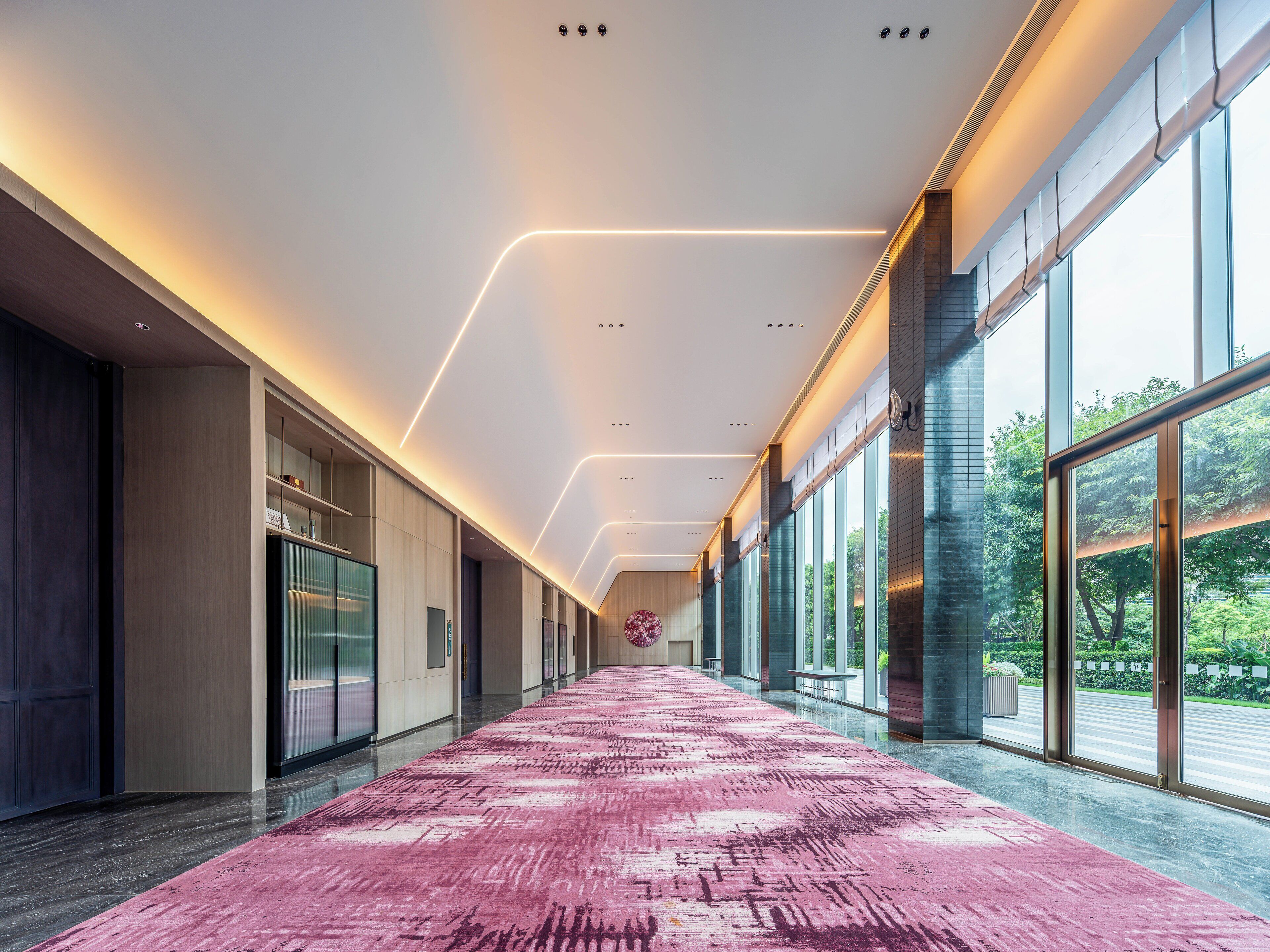 En moderne hotelgang med et langt, pinkfarvet gulvtæppe med mønster, store vinduer med udsigt til grønne områder, og et stilrent, moderne design.