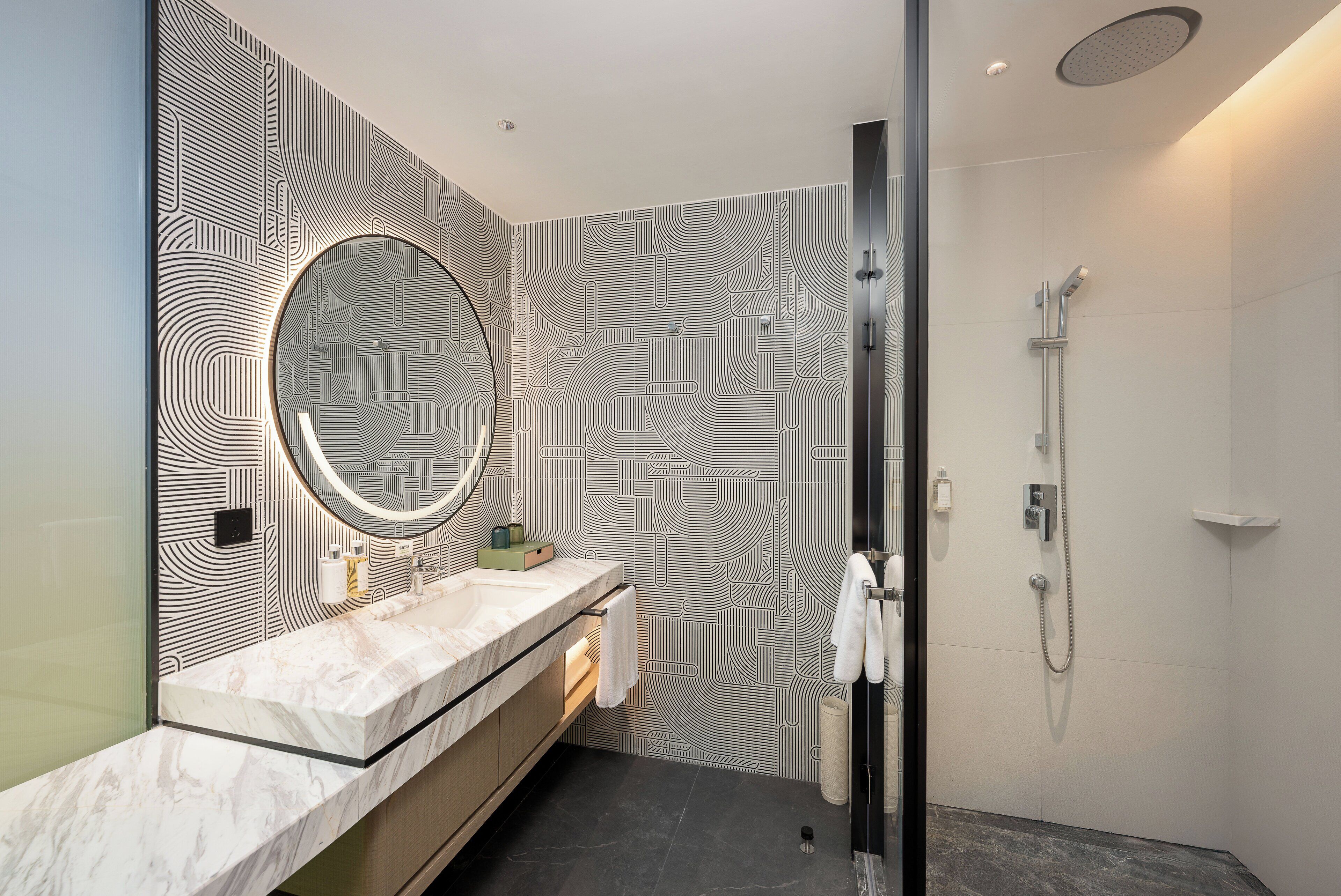 A modern bathroom with a patterned wall, a round mirror, a marble sink, and a shower area.