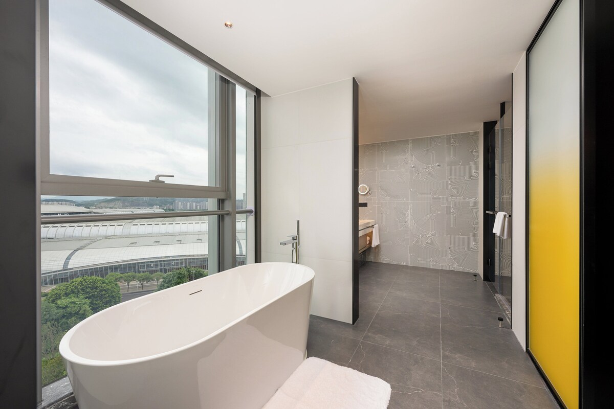 Un baño moderno con una bañera independiente, un ventanal grande y vistas a un paisaje urbano.
