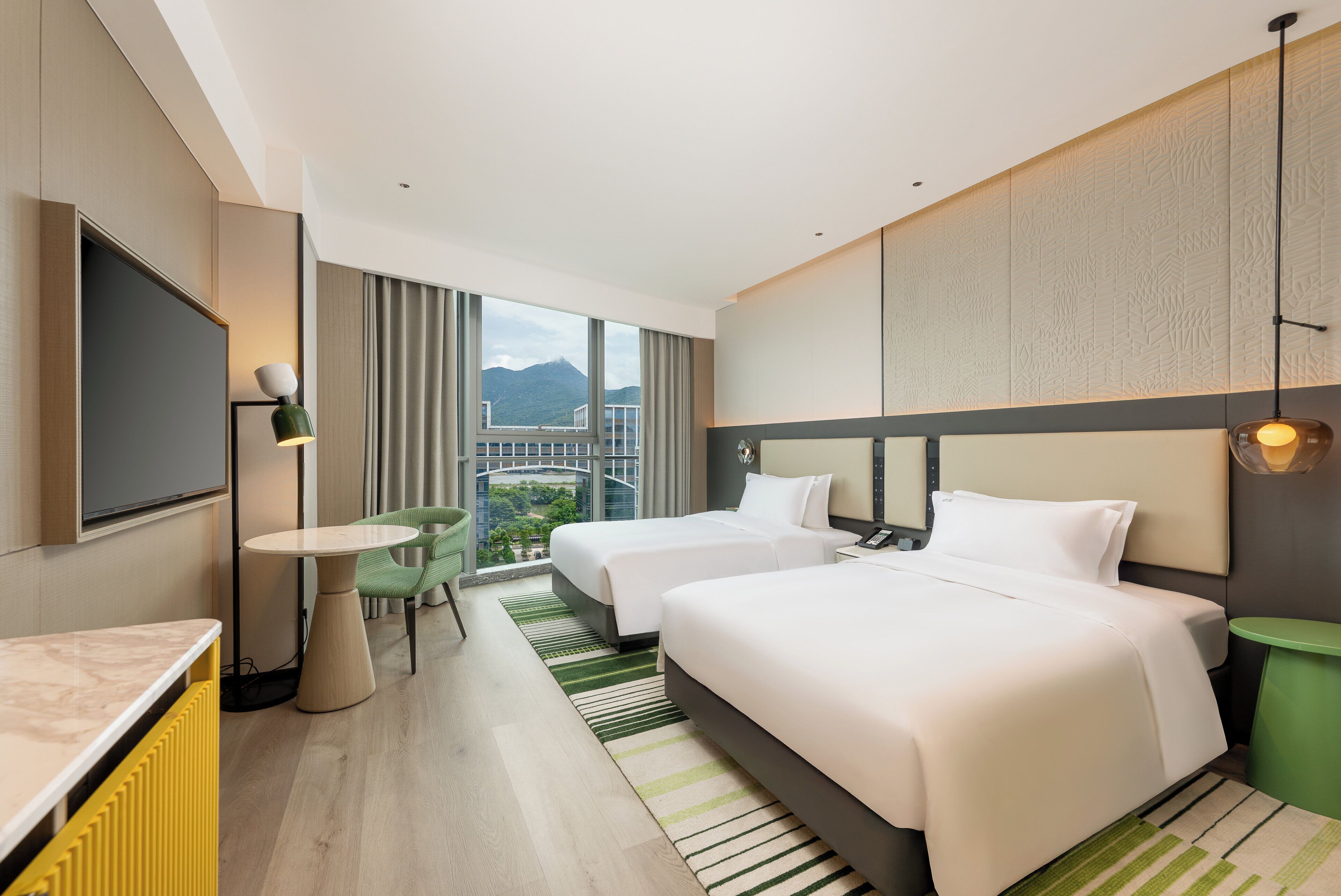 Habitación de hotel con dos camas, un televisor, una mesita y vistas a un paisaje urbano y montañas.