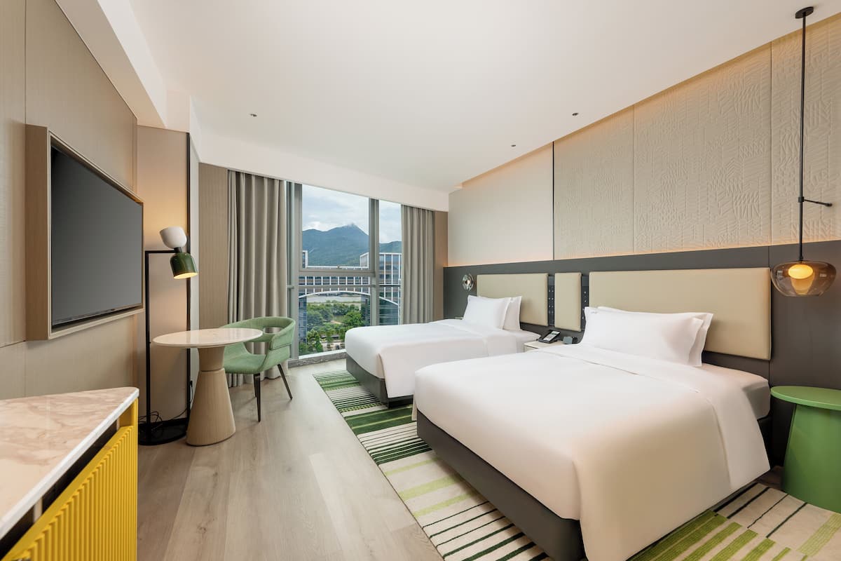 Ein Hotelzimmer mit zwei Betten, einem Fernseher, einem kleinen Tisch und Blick auf eine Stadtlandschaft sowie Berge.