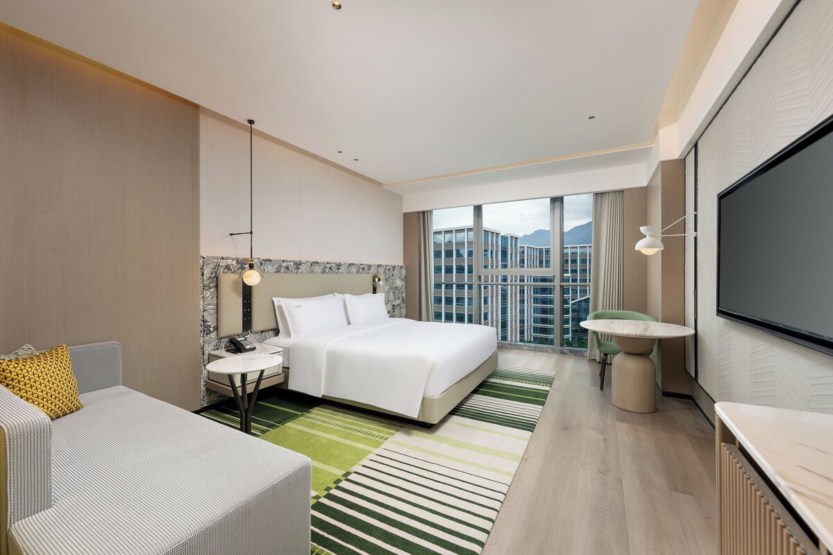 Una habitación de hotel moderna con una cama grande, un área de descanso con una mesa, un televisor y vistas a un paisaje urbano.