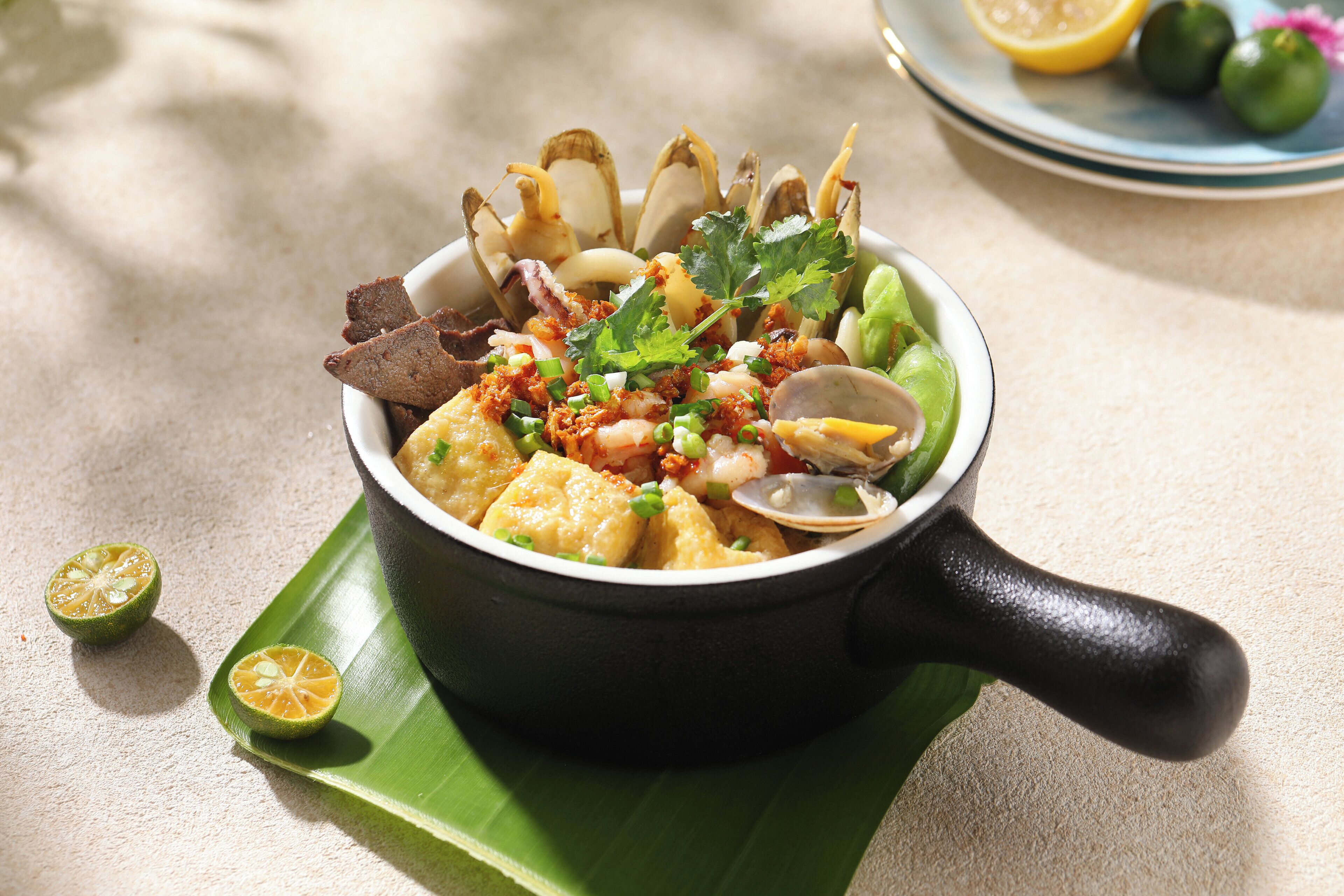 Un tazón de sopa de mariscos con diversos ingredientes, servido en una olla negra con asa, sobre una hoja verde.