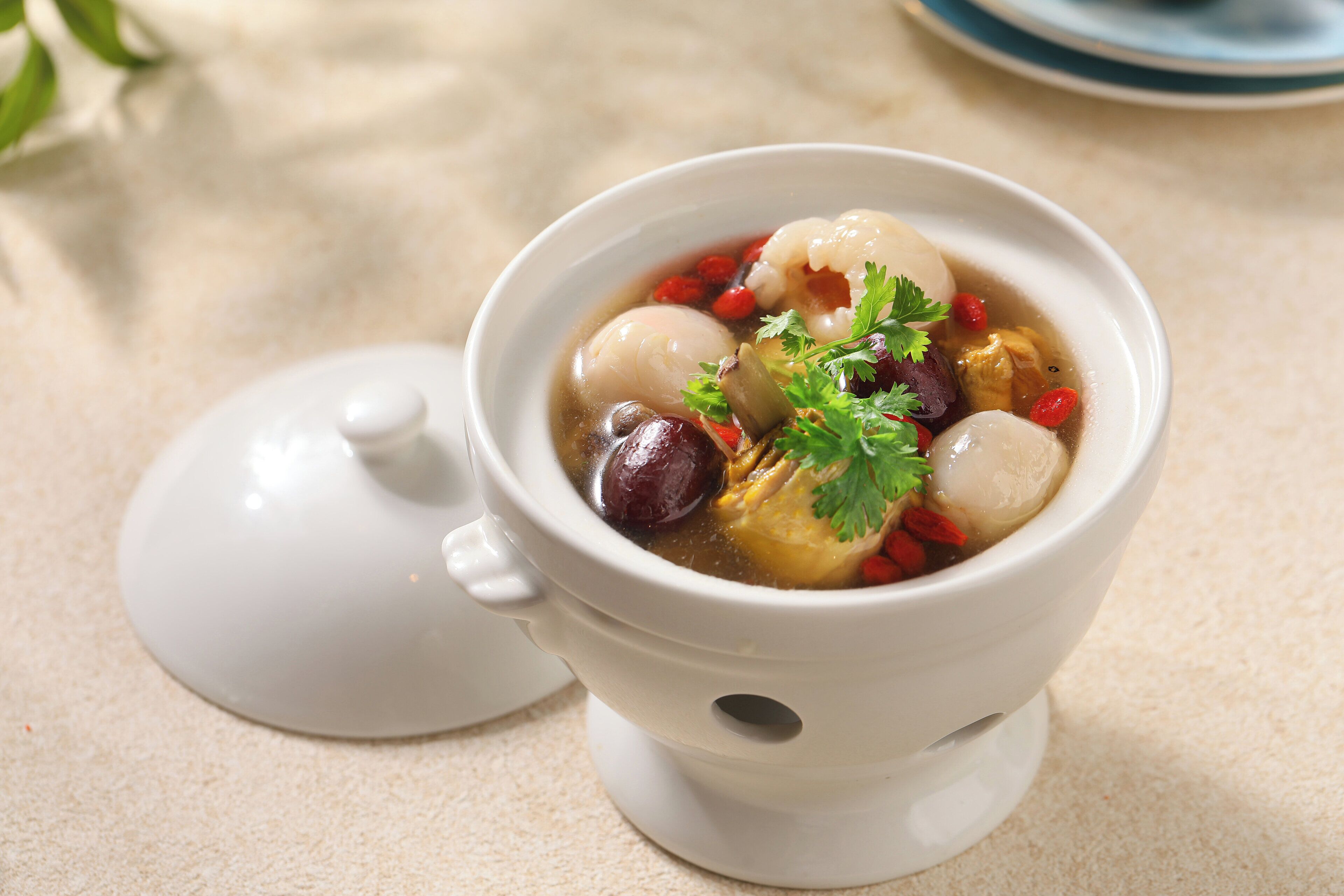 A white ceramic cup with a lid, containing a clear liquid and various ingredients like red dates and goji berries, placed on a beige surface.