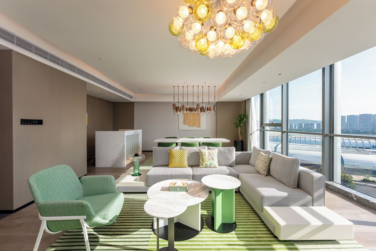Un salon moderne avec un canapé, des fauteuils et une table basse. Une grande fenêtre offre une vue sur la ville.