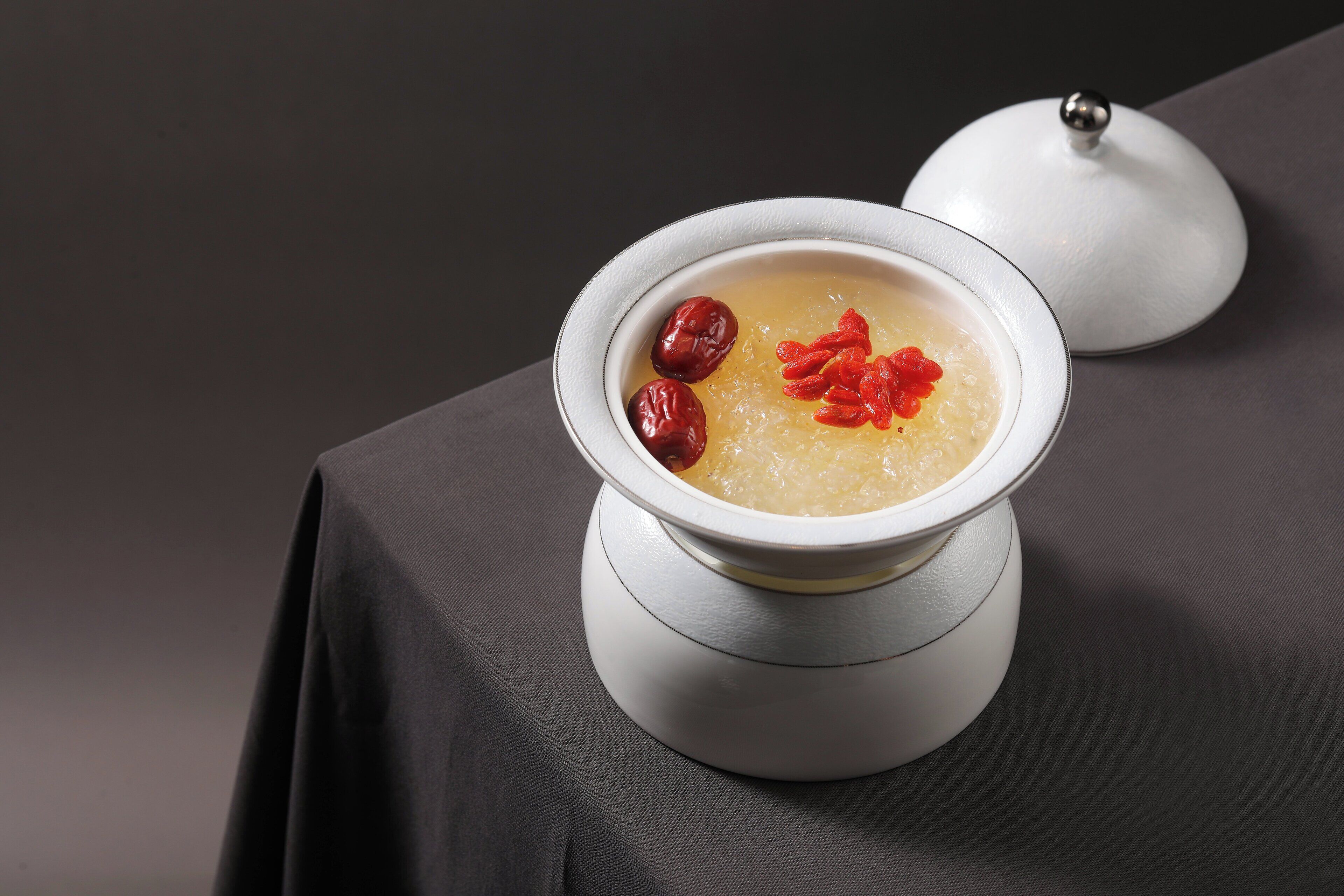 A bowl of soup with red dates and goji berries on a table with a white lid.