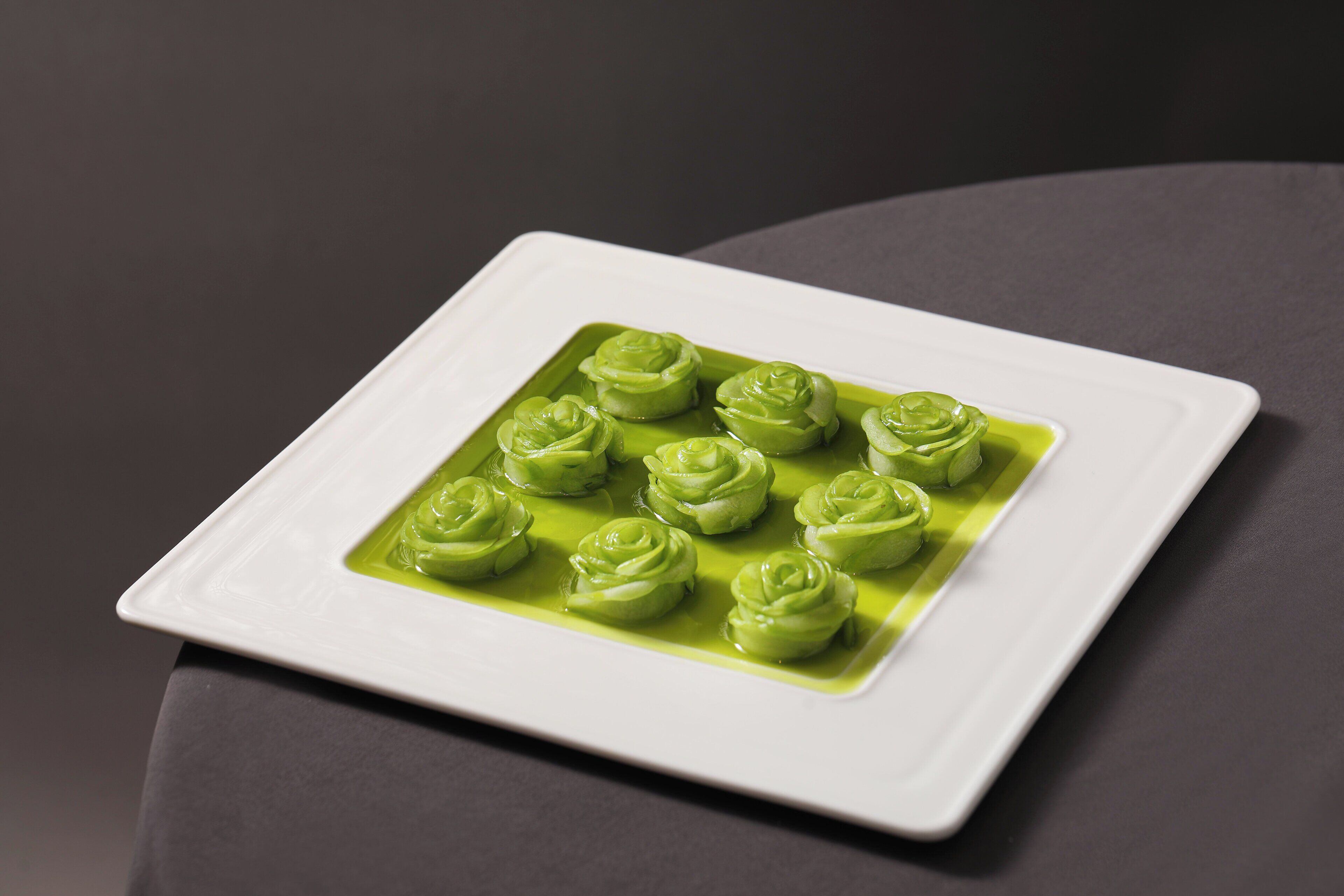 A white square plate with green rose-shaped food items on a dark surface.