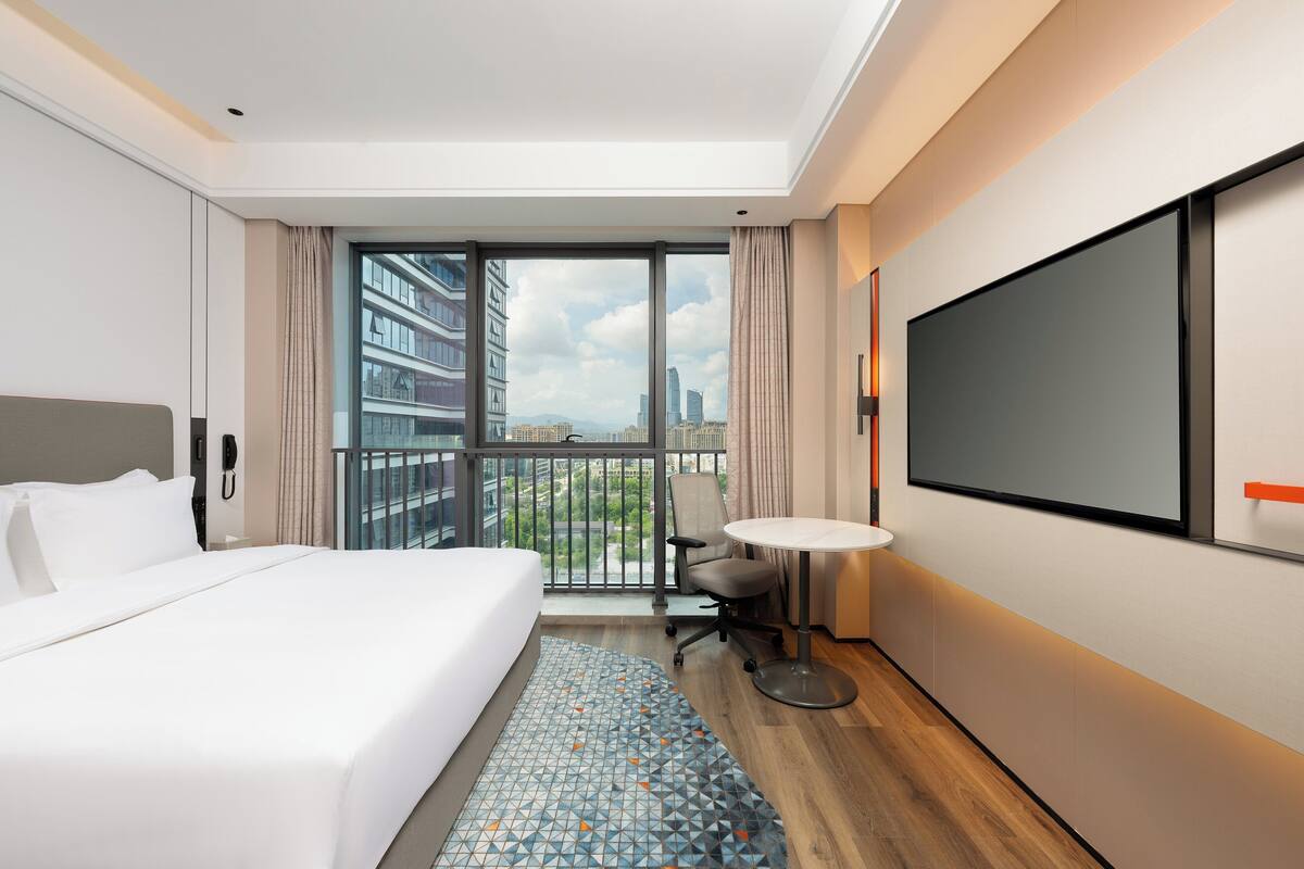 A hotel room with a large bed, a desk with a chair, a TV, and a balcony with a city view.