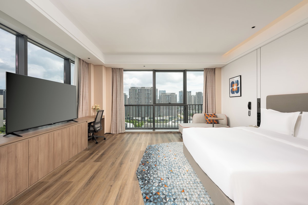 Habitación de hotel moderna con una cama grande, un escritorio con televisión de pantalla plana, una silla y vistas a la ciudad a través de amplios ventanales.